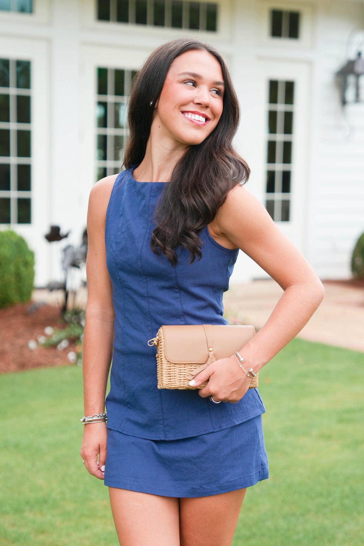 Woman in a blue dress holding a beige clutch in front of a house.