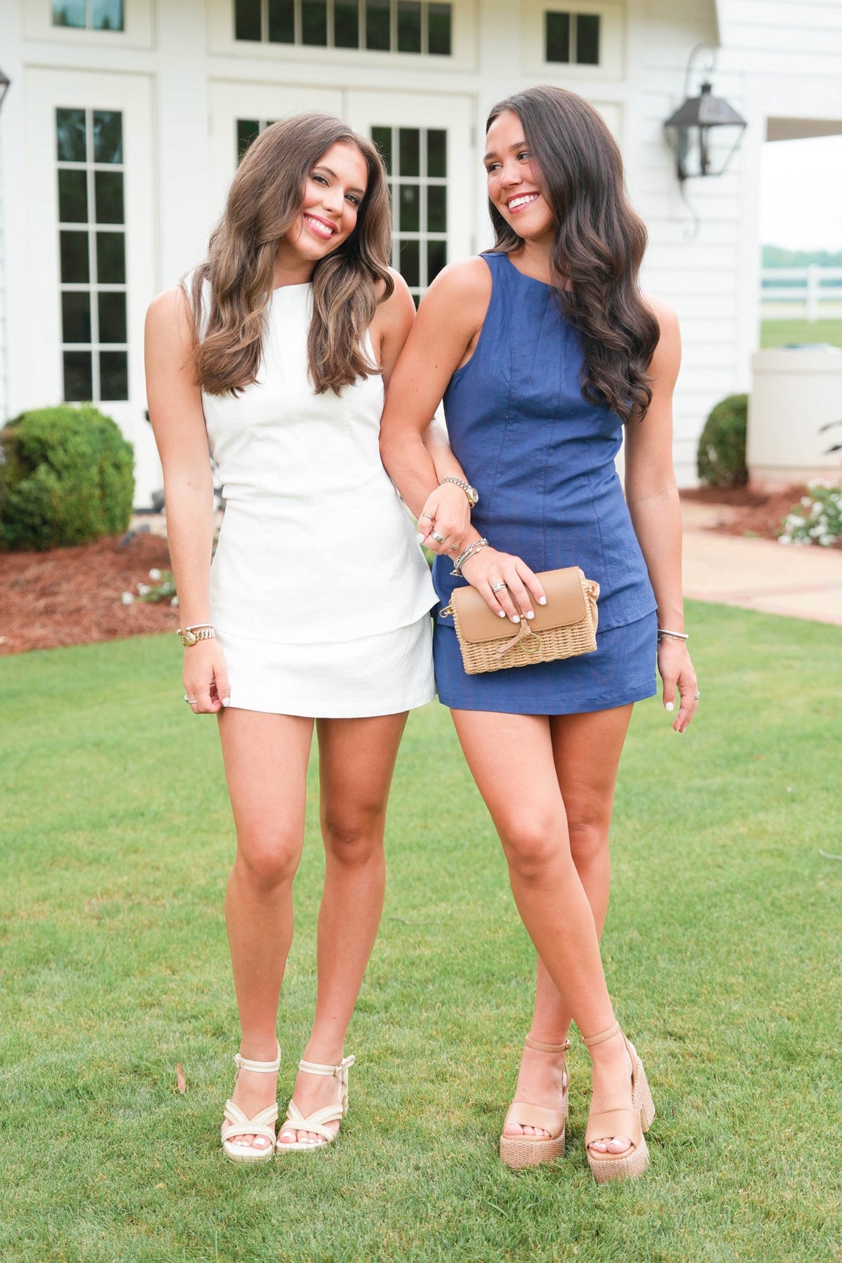 Two women standing on grass wearing dresses with a house in the background