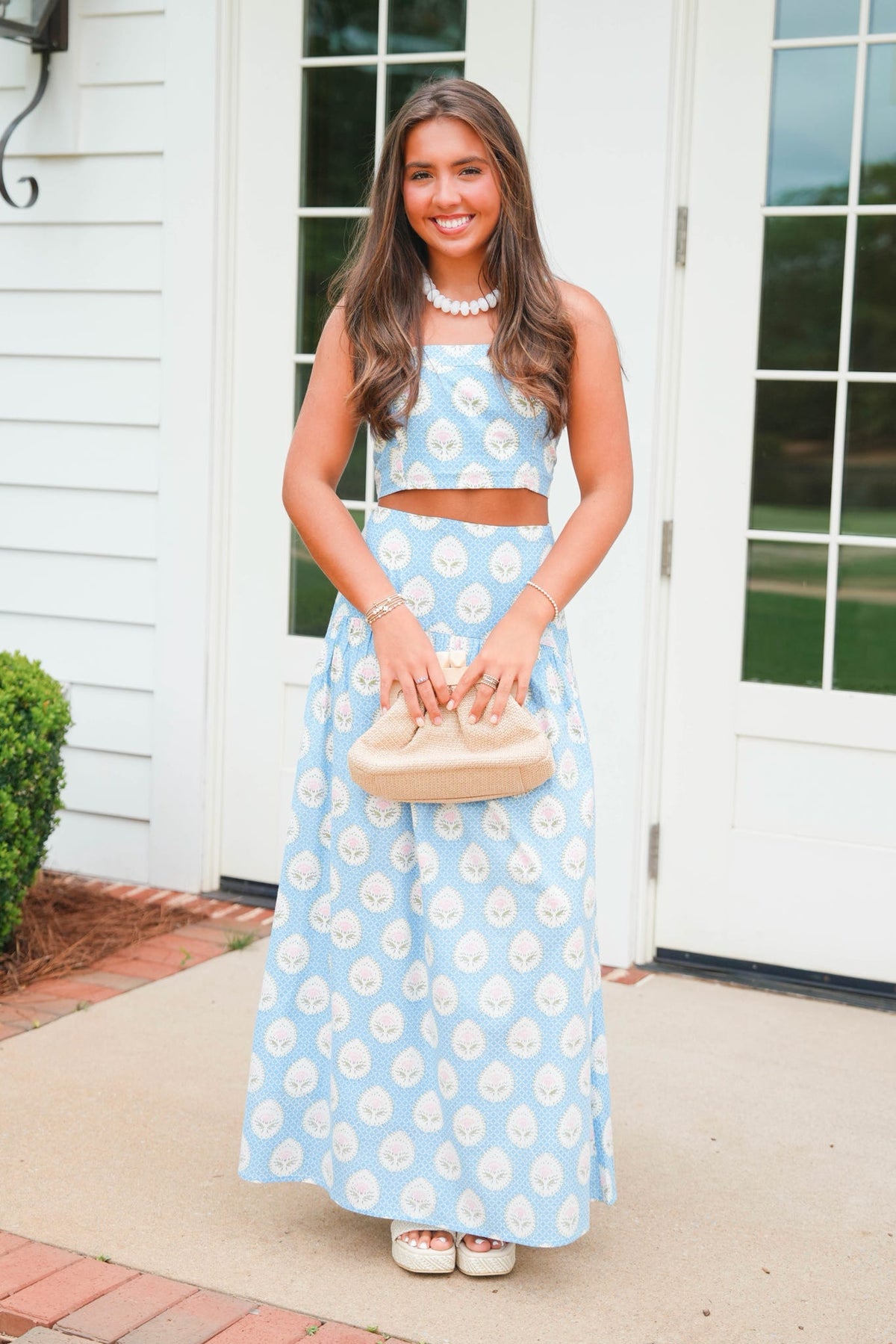 Woman in a blue floral dress holding a beige handbag outdoors.