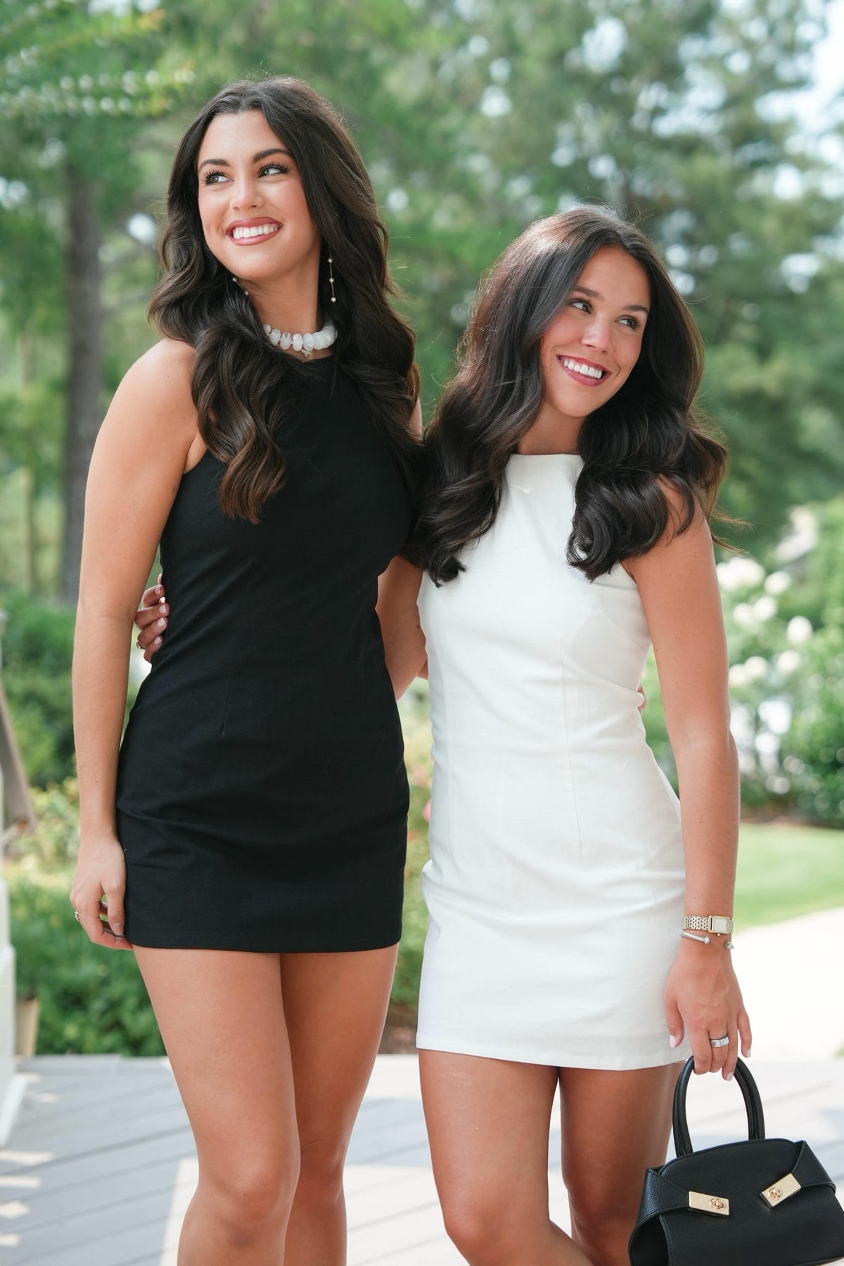 Two women in black and white dresses standing outdoors with greenery in the background