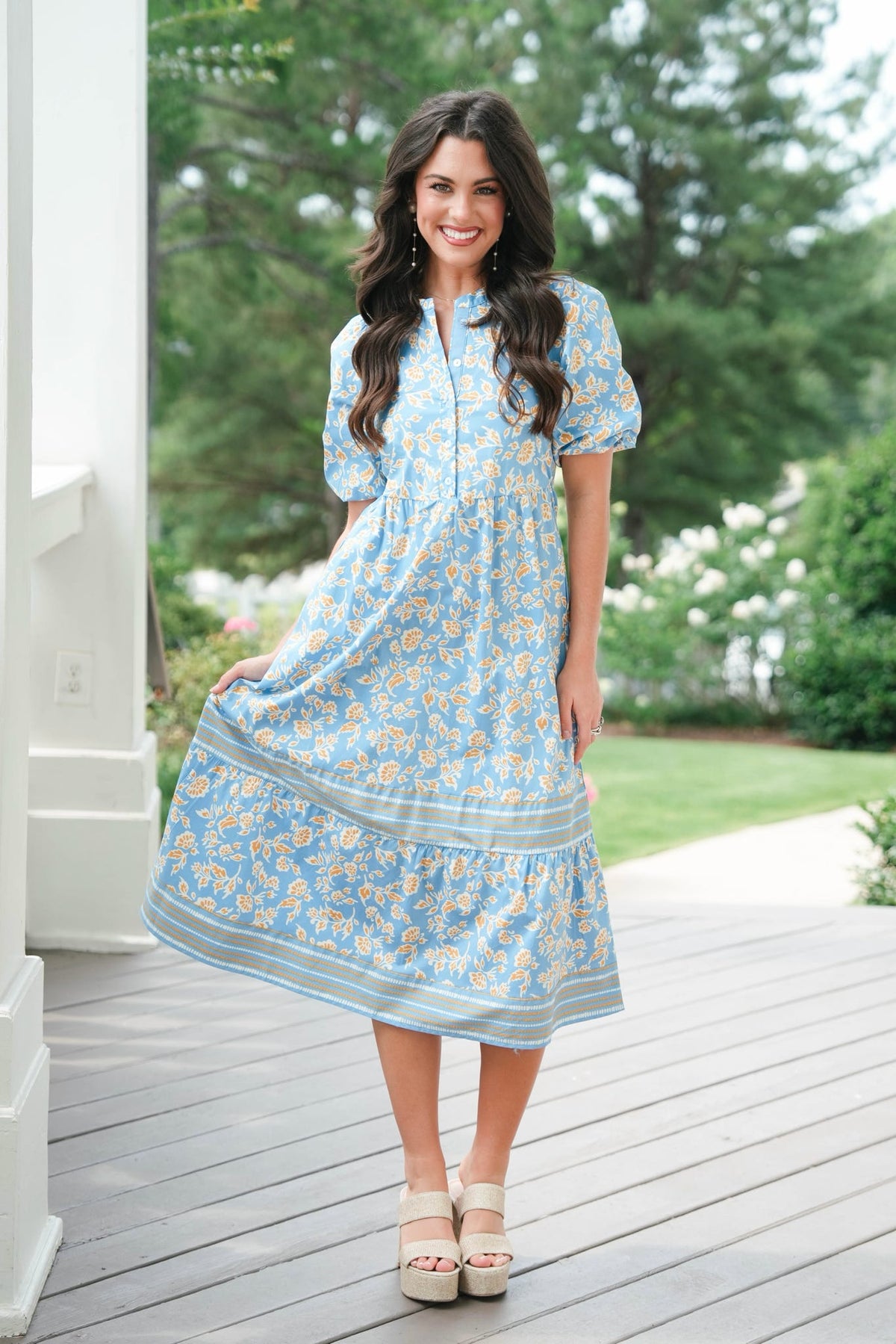 Woman in a blue floral dress standing on a wooden deck with greenery in the background