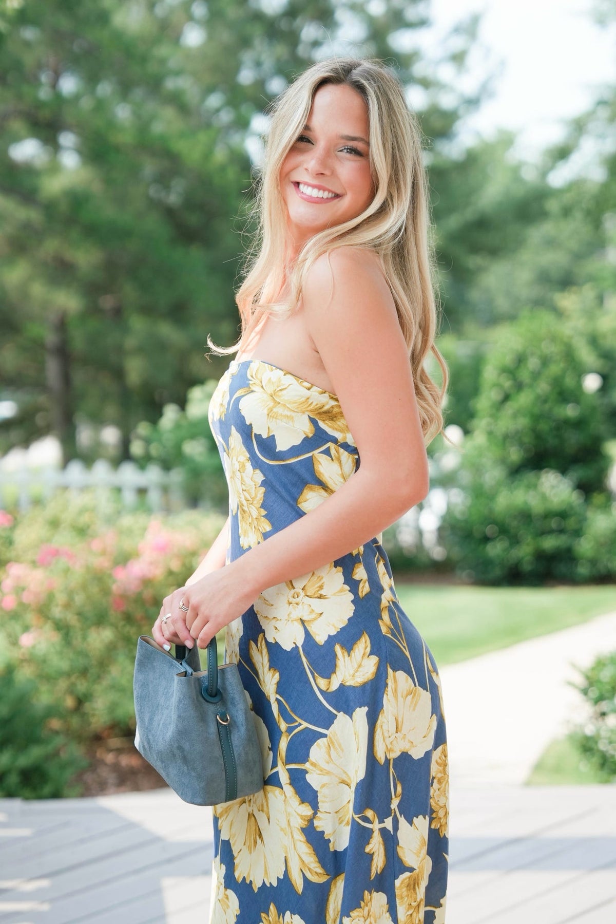 Woman in a blue floral dress holding a small blue handbag outdoors.