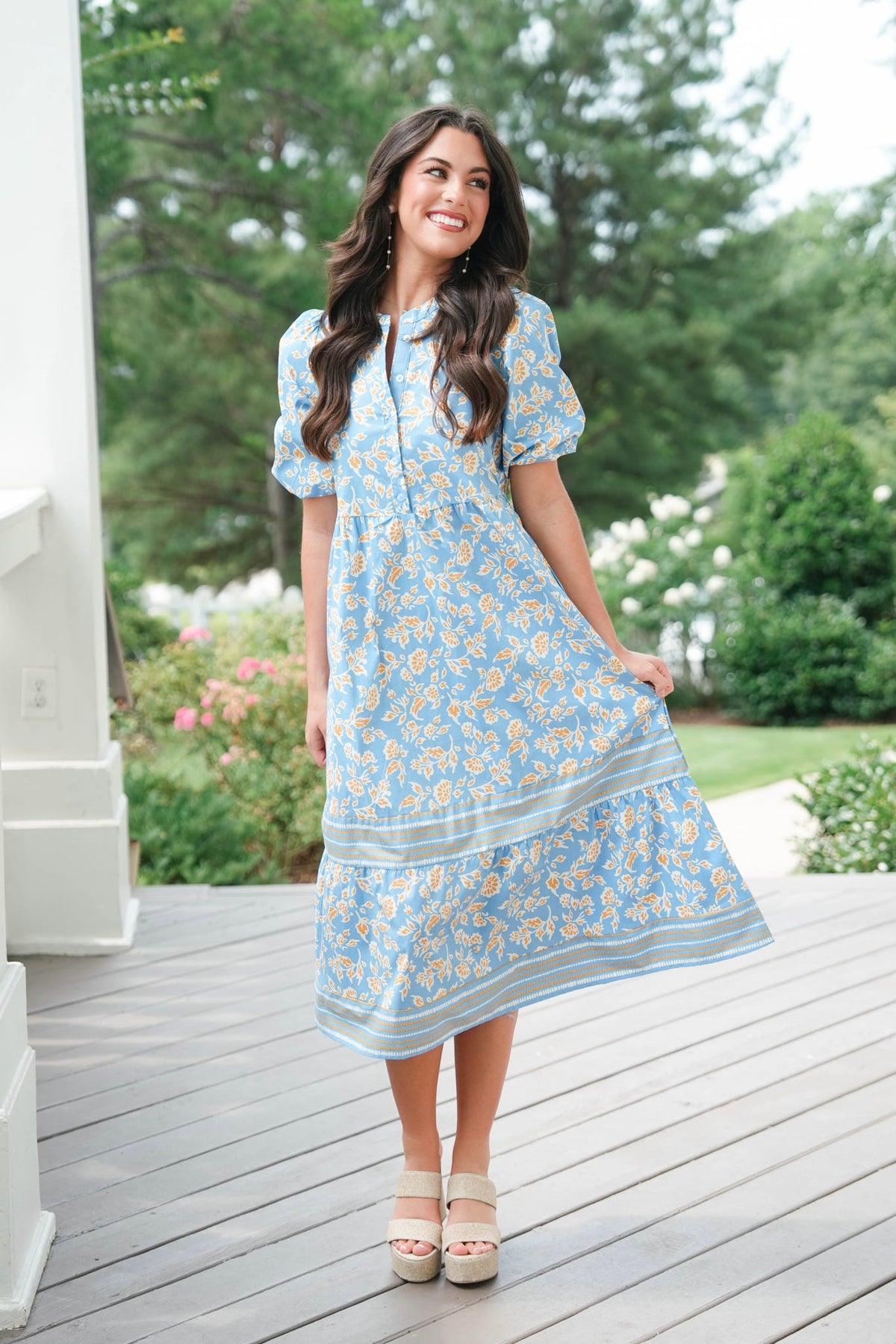 Woman in a blue floral dress standing on a wooden deck with greenery in the background