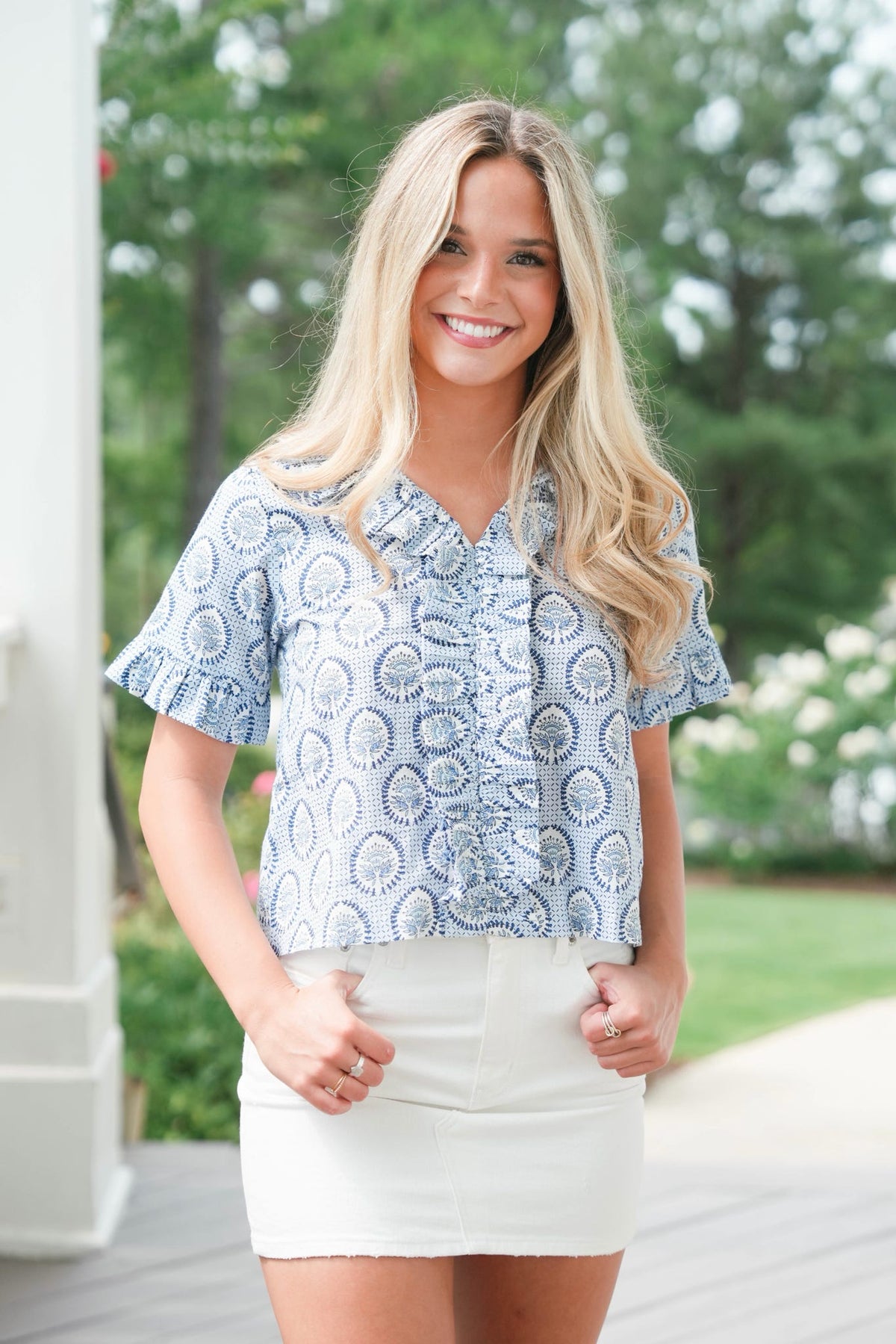 Woman wearing a blue patterned blouse and white skirt outdoors with greenery in the background
