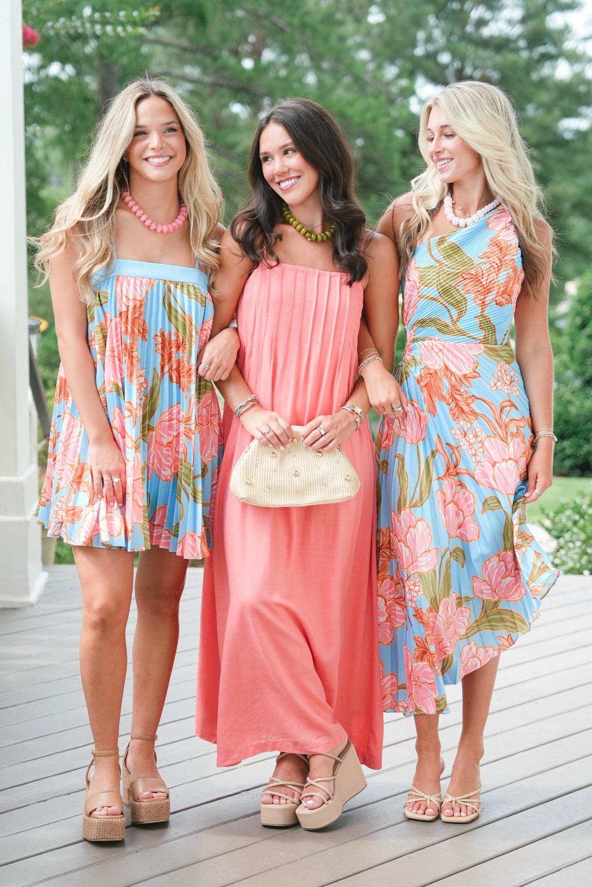 Three women in floral dresses standing on a wooden deck with greenery in the background
