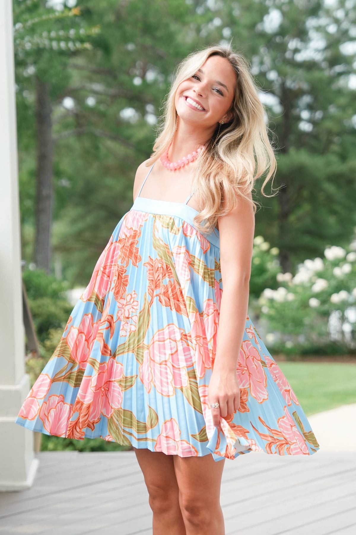 Woman wearing a colorful floral dress standing on a wooden deck with greenery in the background