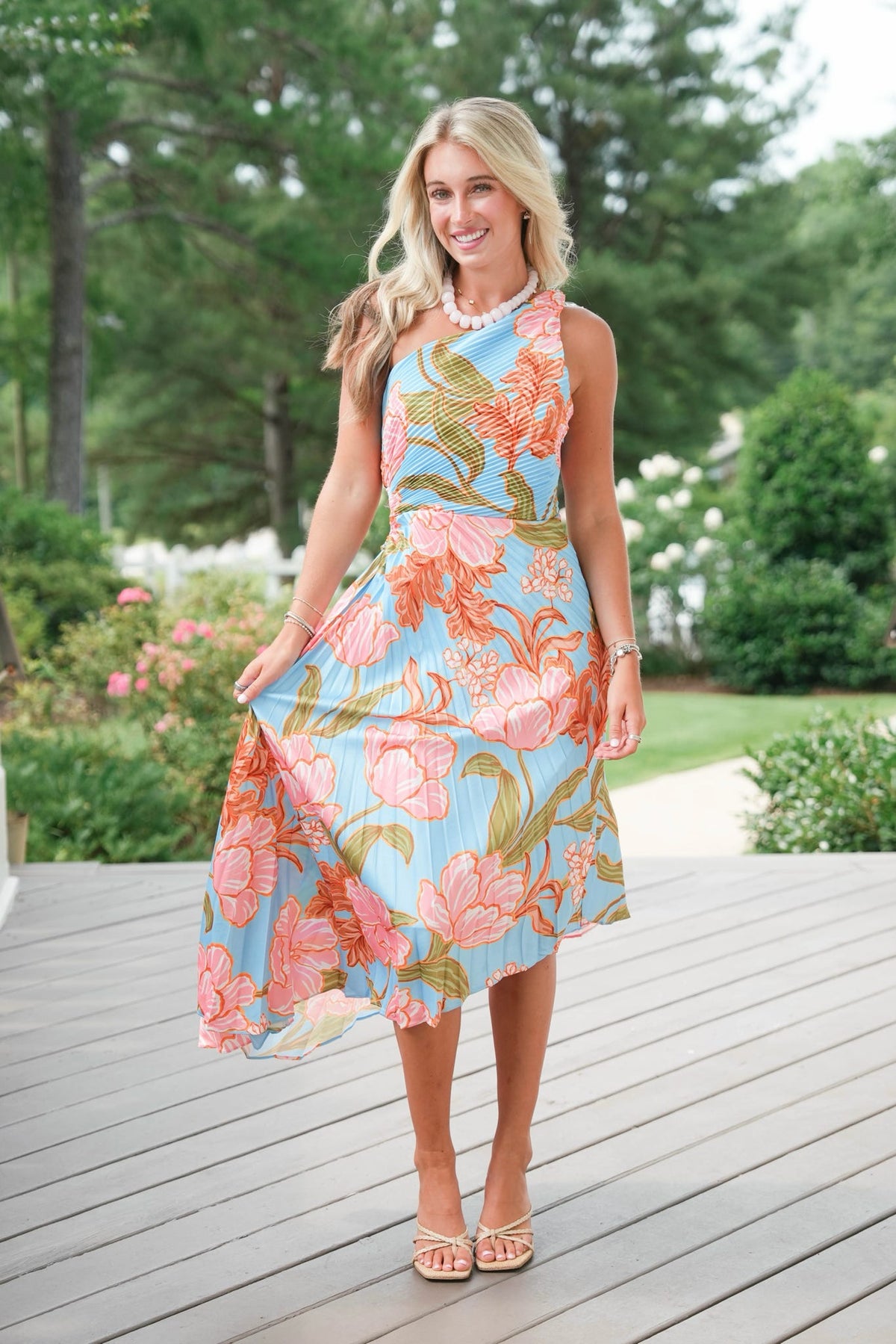 Woman wearing a floral dress standing on a wooden deck with greenery in the background