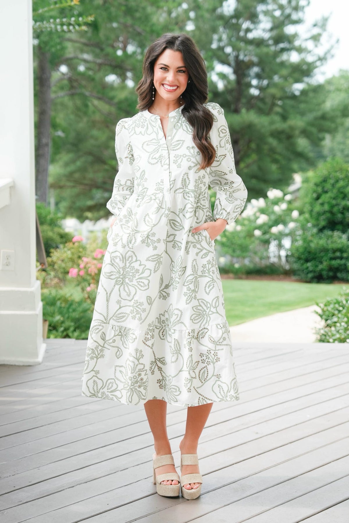 Woman in a white floral dress standing on a wooden deck with greenery in the background
