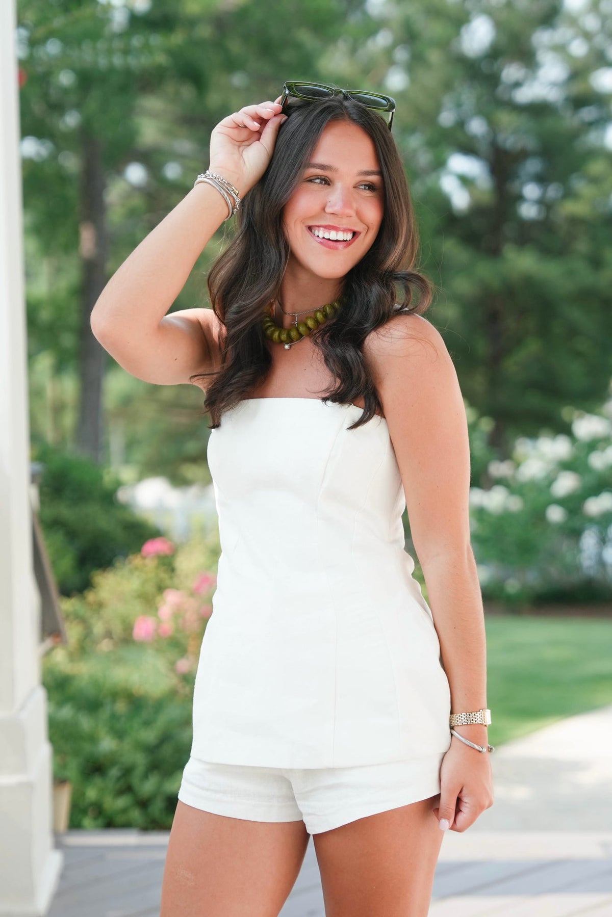 Woman in a white strapless top and shorts standing outdoors with greenery in the background