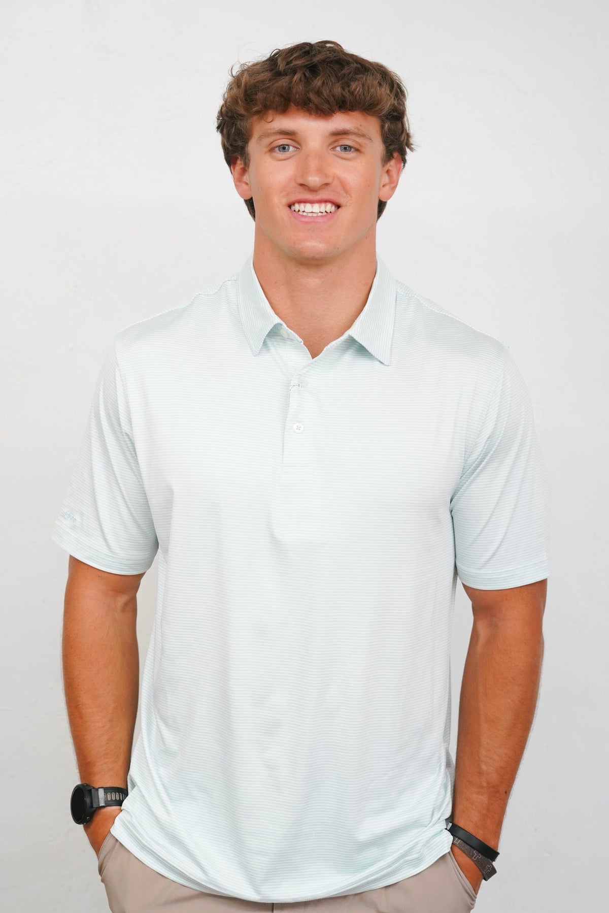 A smiling young man with curly brown hair wearing a light mint green short-sleeve polo shirt and khaki pants, standing against a plain light gray background.
