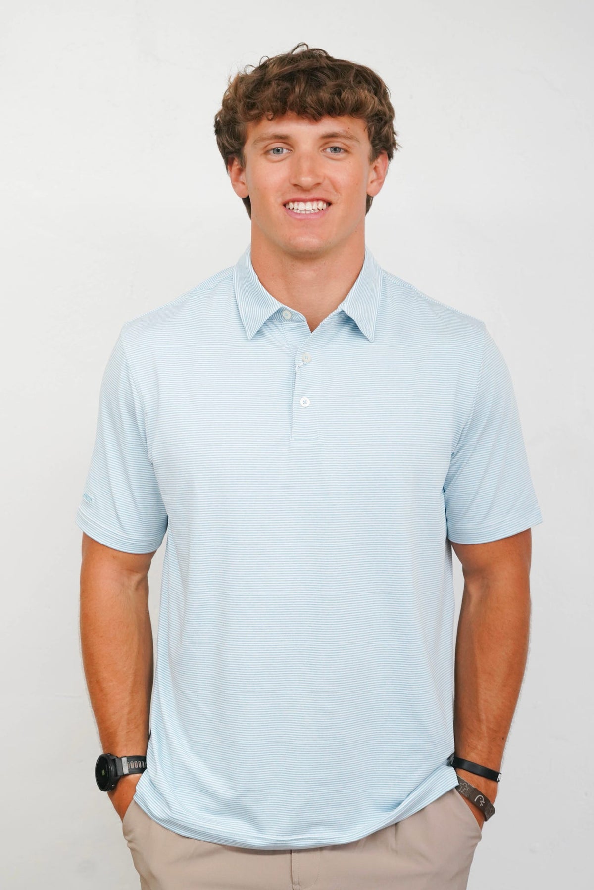 A smiling young man with curly brown hair wearing a light blue short-sleeve polo shirt and khaki pants, standing against a plain light gray studio background.