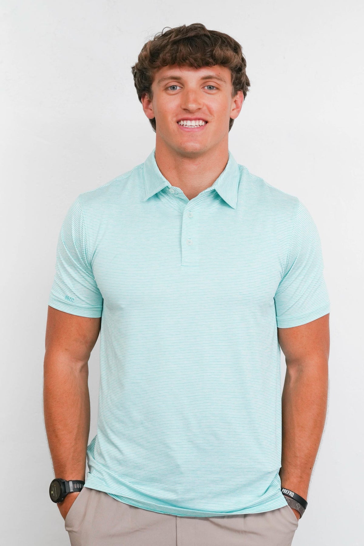 A smiling man wearing a light mint green short-sleeve polo shirt and khaki pants, standing against a plain light gray background.
