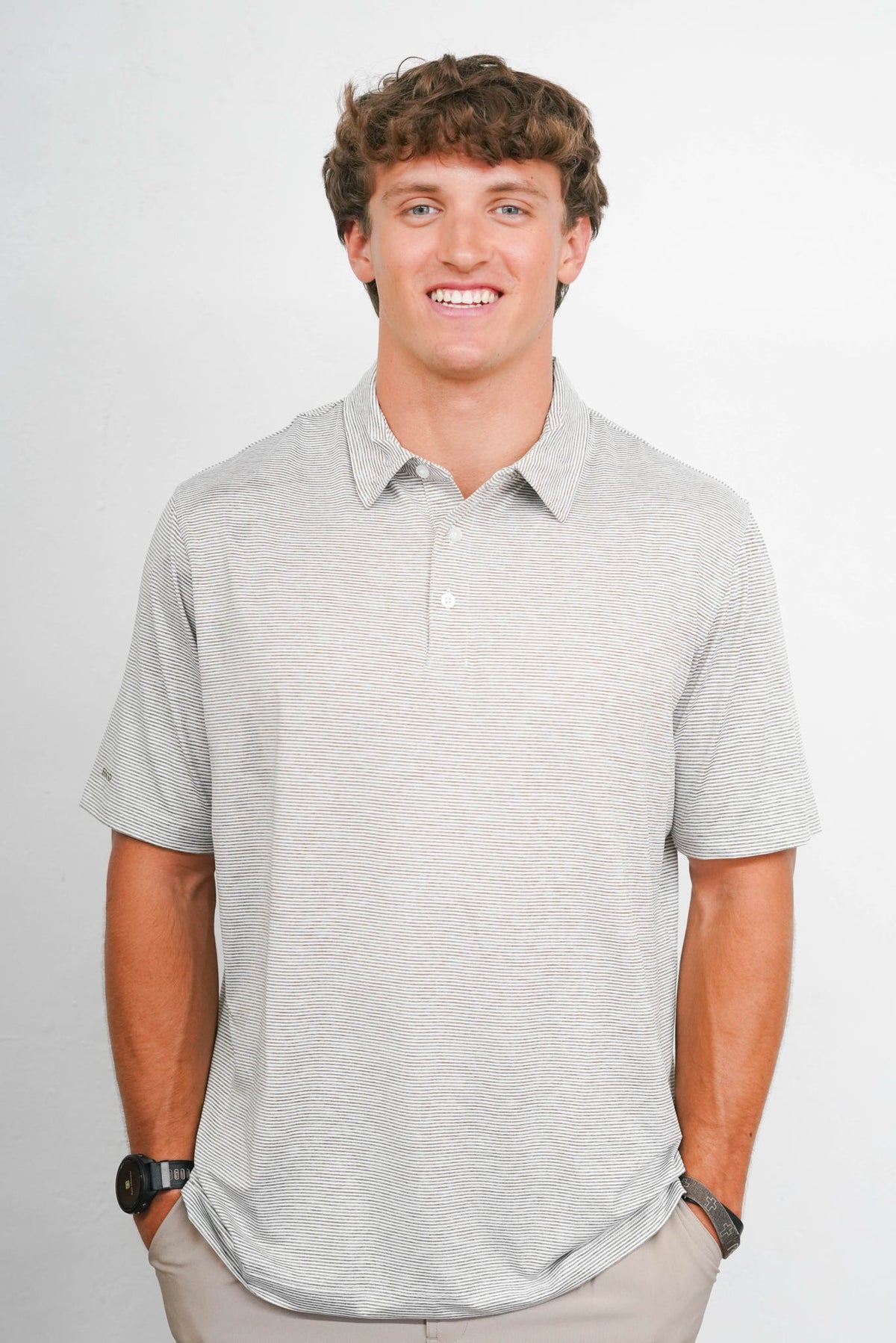 A smiling young man with curly brown hair wearing a light heather gray short-sleeve polo shirt, khaki pants, and a black watch, standing against a light gray studio background.