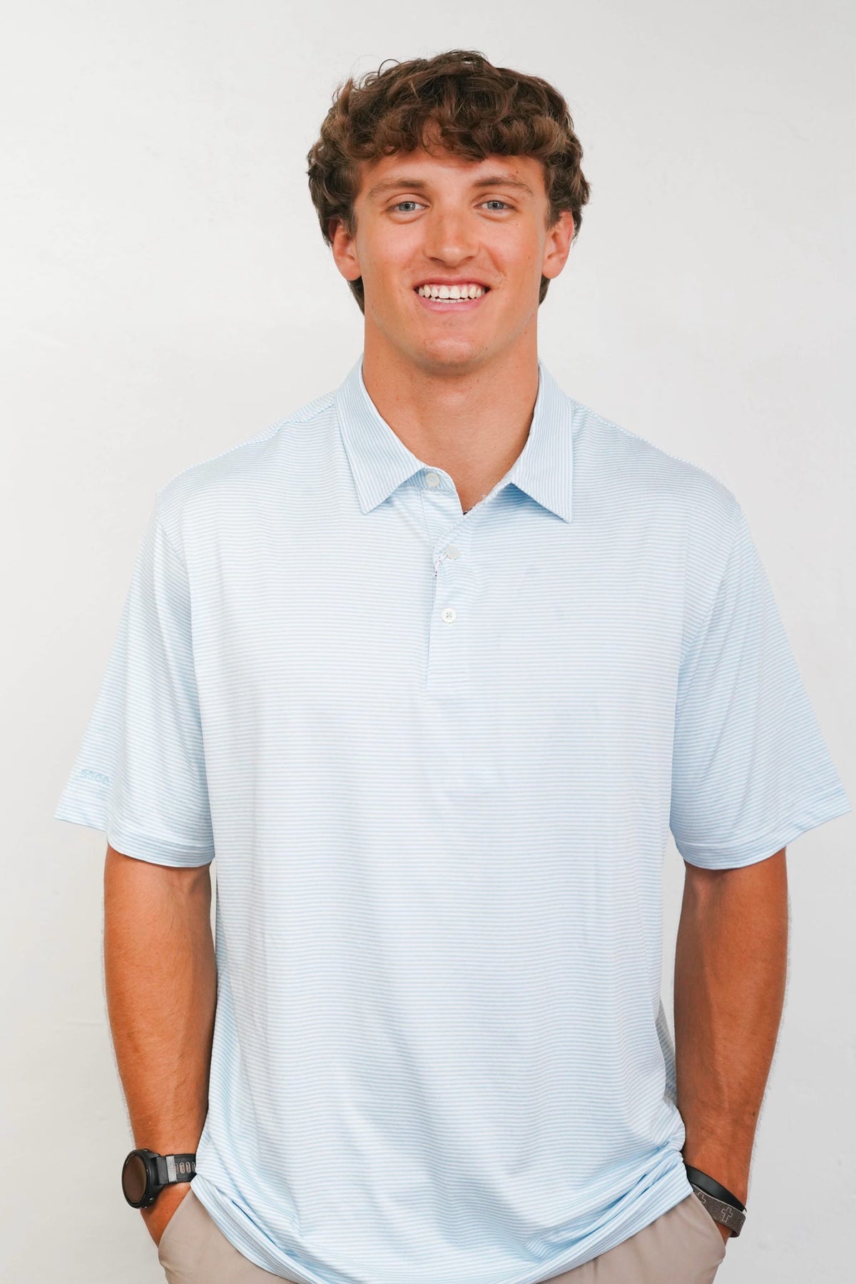 A smiling young man with curly brown hair wearing a light blue short-sleeve polo shirt, standing against a plain light gray studio background.