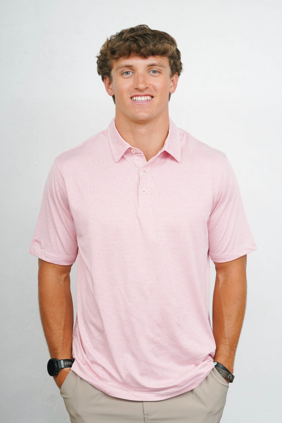 A smiling man wearing a light pink short-sleeve polo shirt and khaki pants, standing against a light gray studio background.