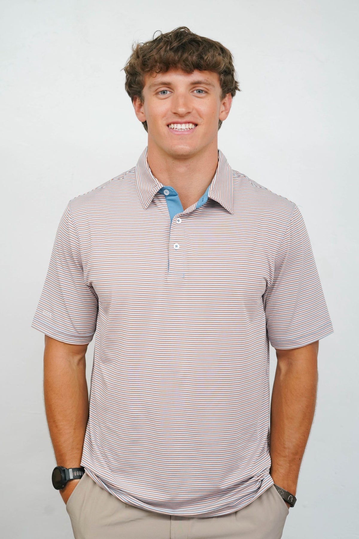A smiling man wearing a short-sleeve polo shirt with fine horizontal stripes in beige and light blue, paired with khaki pants, photographed against a plain light gray background.