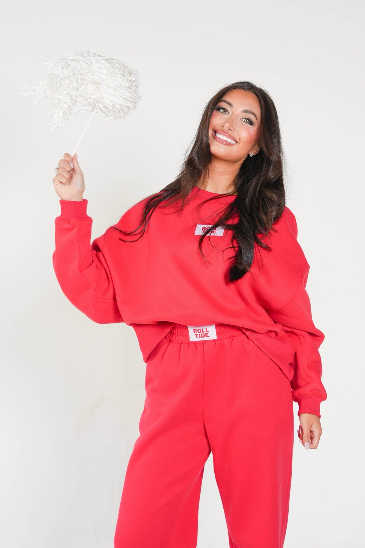 A woman modeling a bright red sweatshirt and matching sweatpants set against a plain white studio background, holding a white decorative pom-pom.