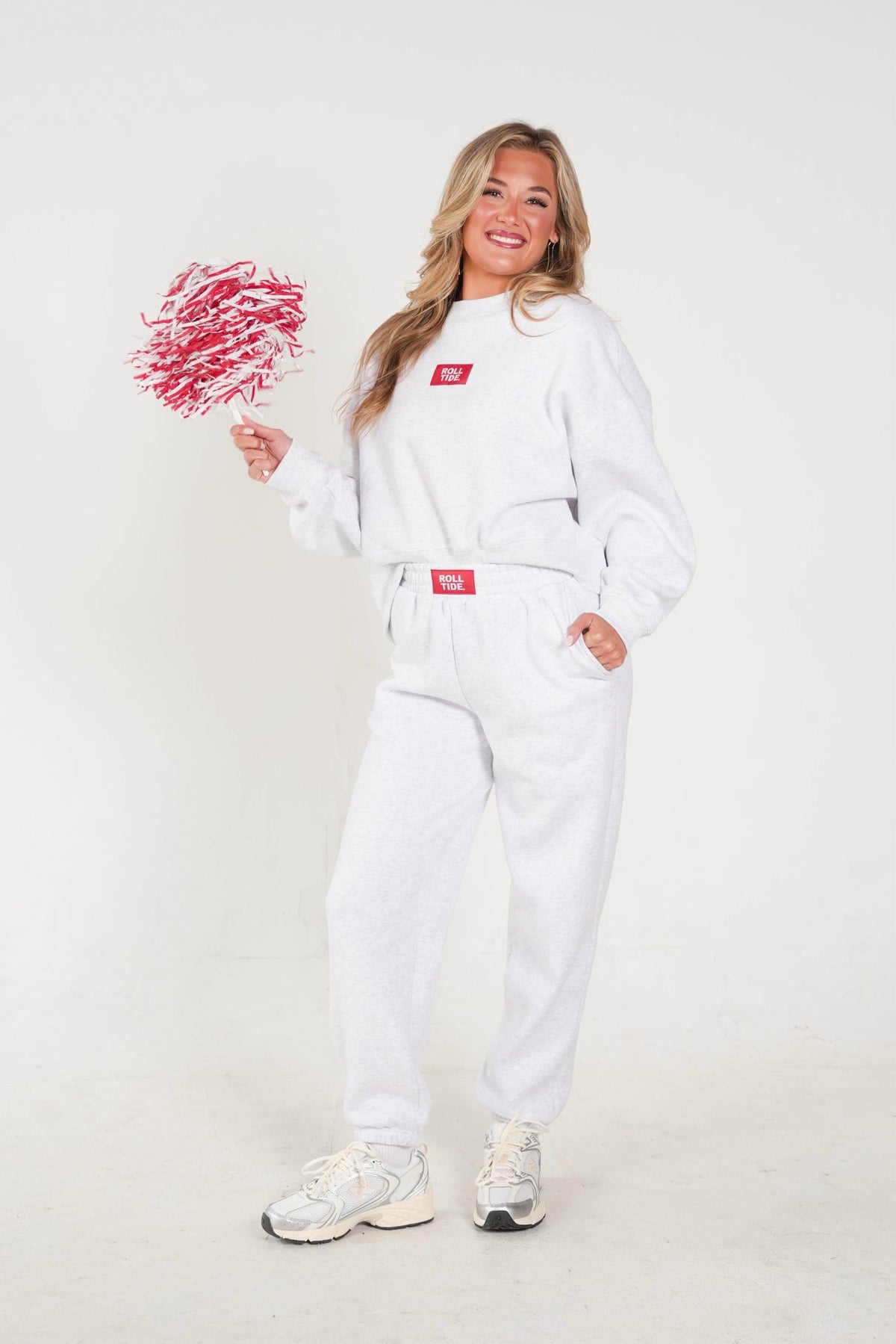 A woman modeling a white sweatshirt and matching sweatpants set with red accents, holding a red scribble-shaped prop against a plain light background.