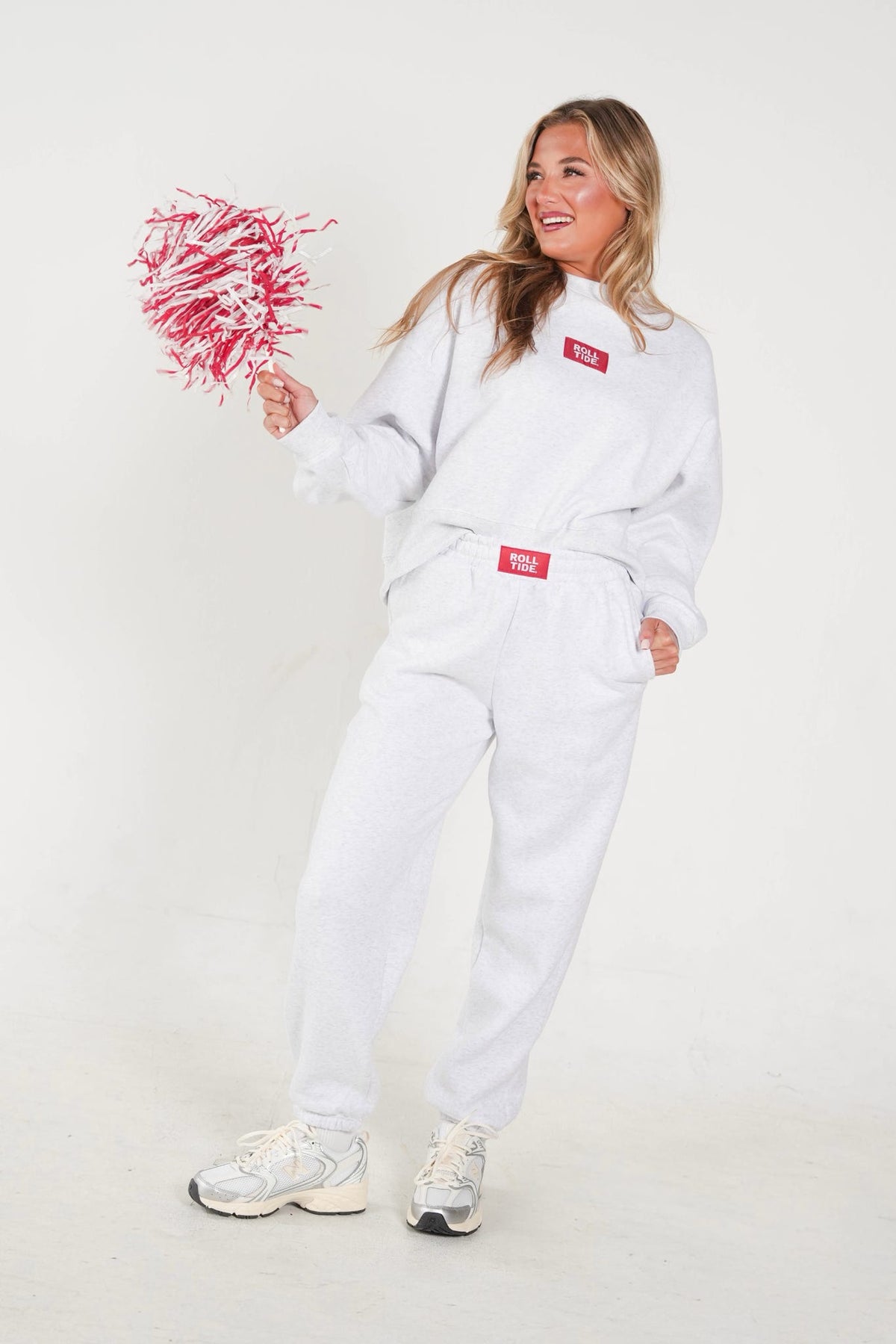 A woman modeling a white two-piece tracksuit with red rectangular logo patches on the chest and waist, holding a red and white pom-pom against a plain white studio background.