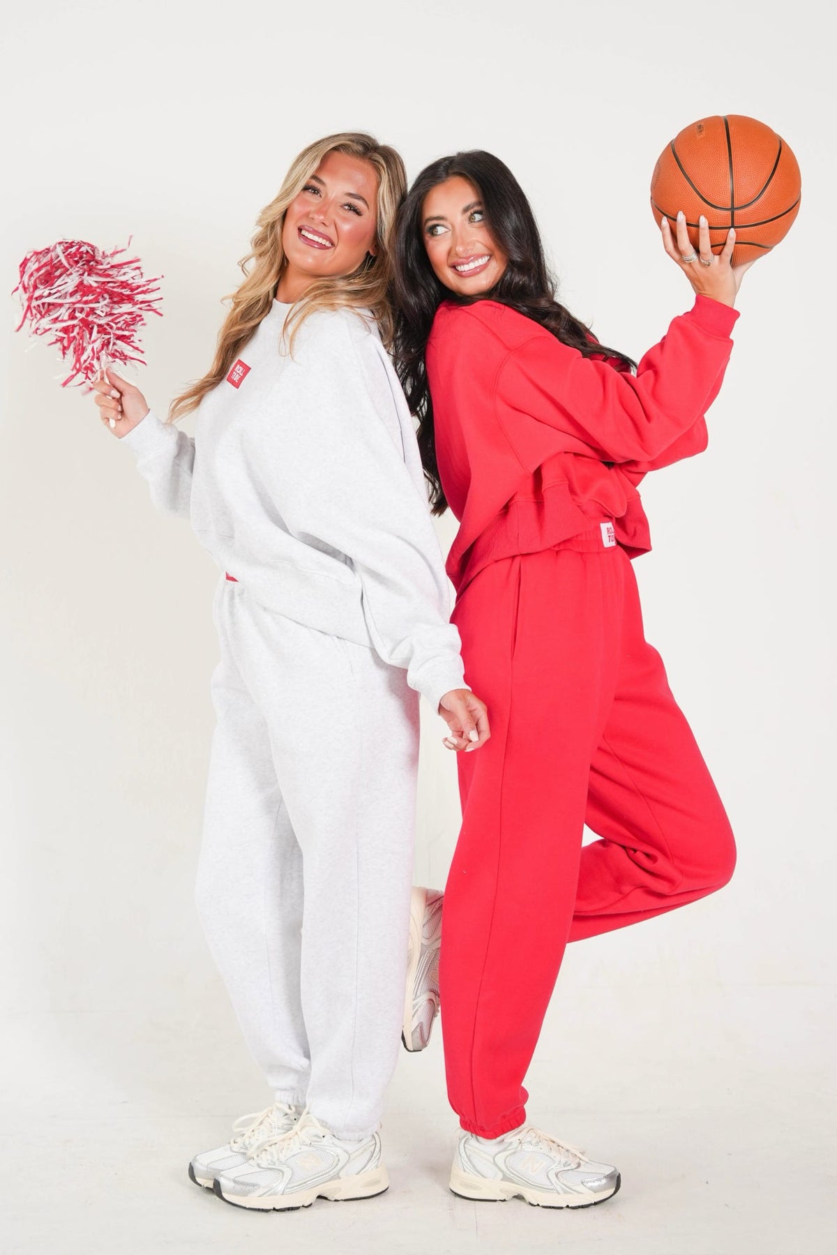 Two women posing in matching athletic tracksuits, one in a white sweatshirt and joggers holding a pink pom-pom, and the other in a red sweatshirt and joggers holding an orange basketball, against a plain white studio background.