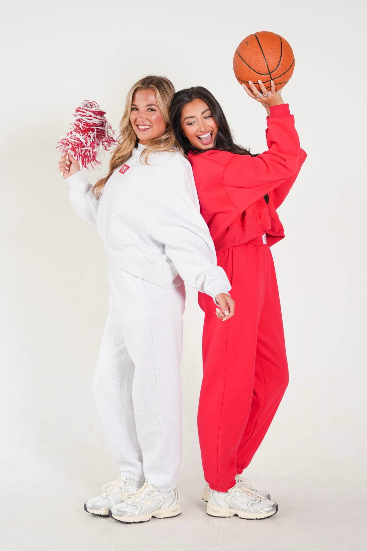 Two women posing in matching sweatshirt and jogger tracksuits, one in white and one in bright red, holding a basketball and a pink and white pom-pom against a plain white studio background.