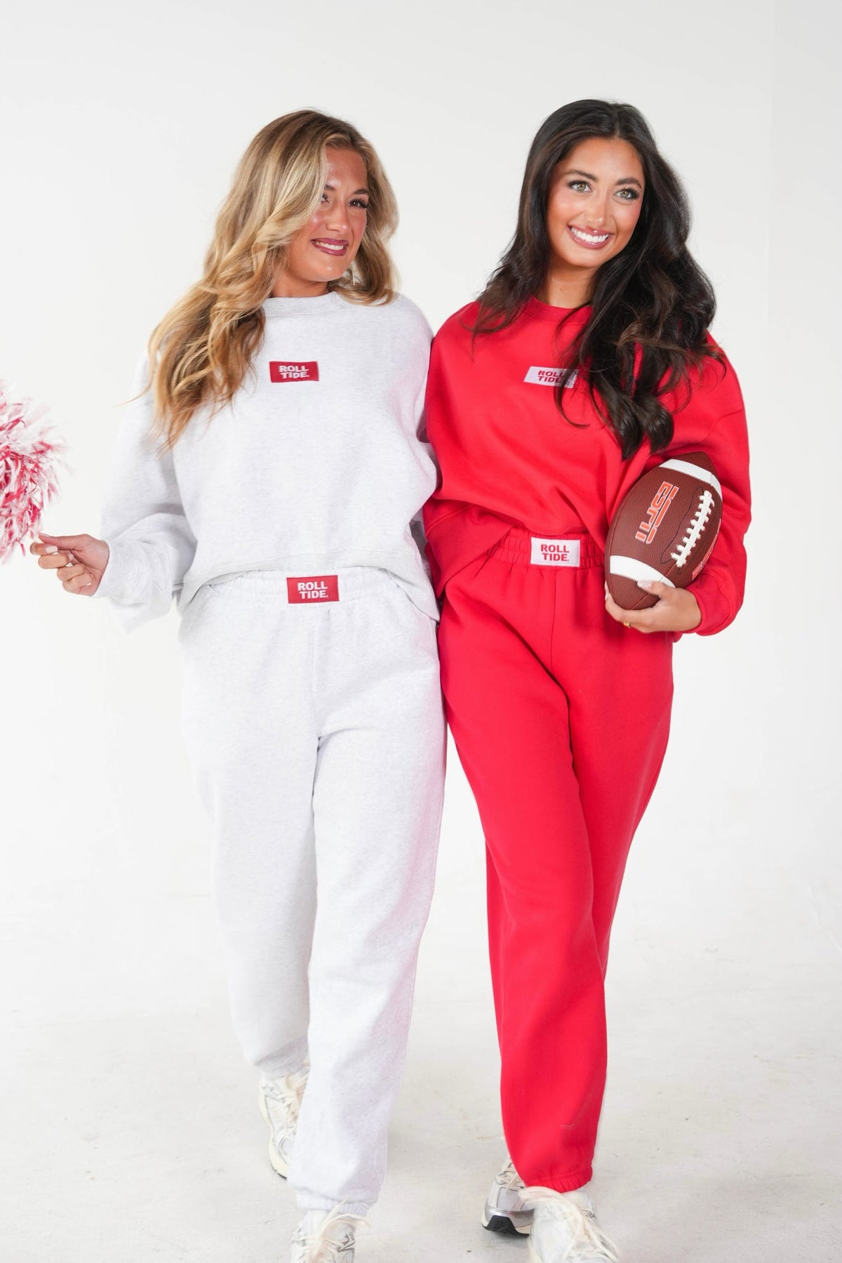 Two women modeling matching sweatshirt and jogger sets in white and red, each with a small red rectangular chest patch. The woman on the right holds a brown football, and the background is a clean white studio backdrop.