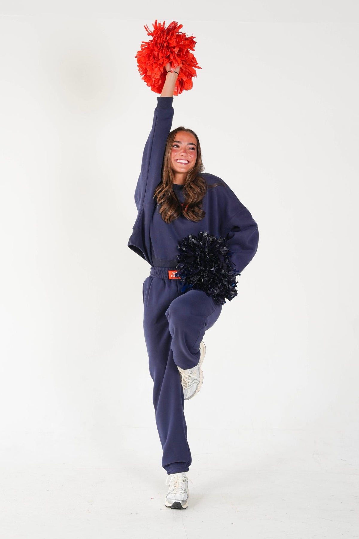 A woman in a navy blue sweatshirt and sweatpants jumps while holding red and black pom-poms against a plain white studio background.