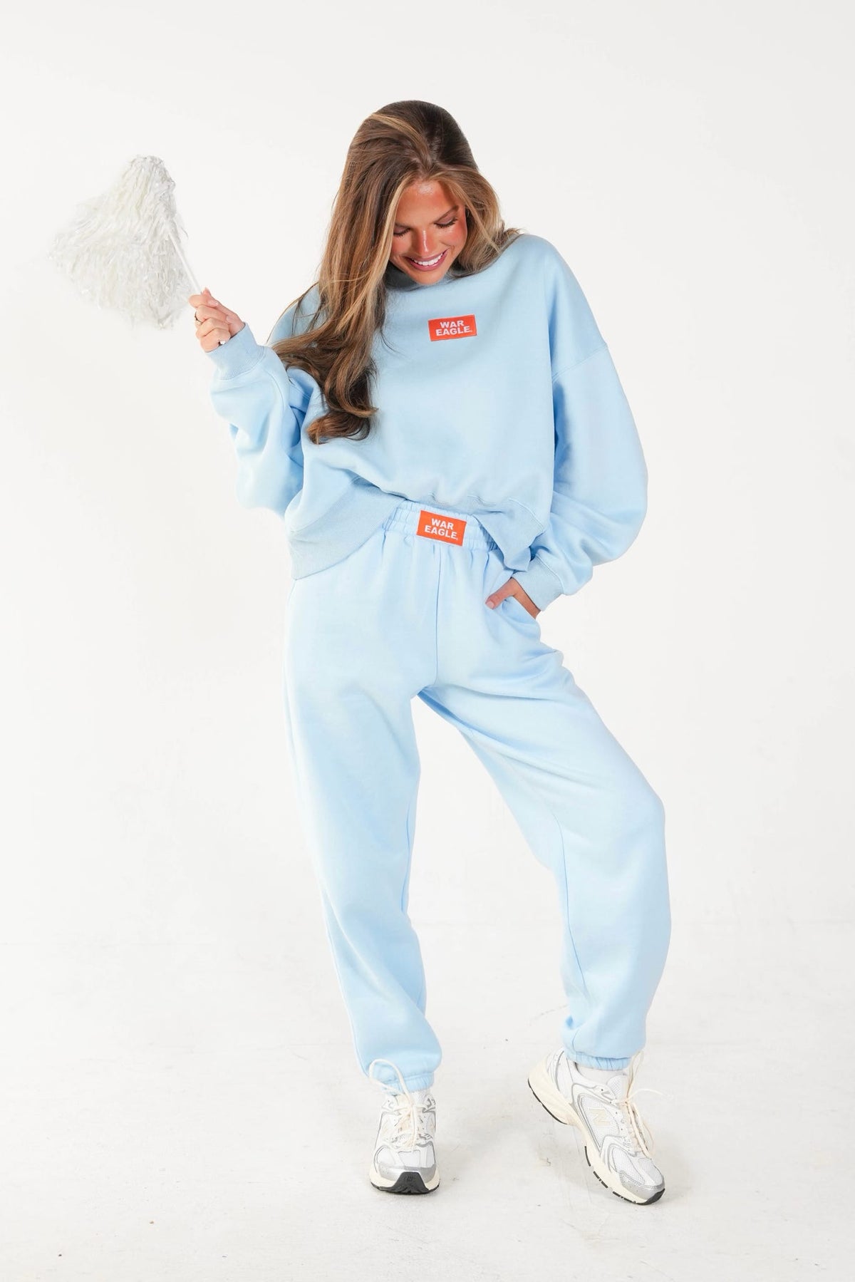 A woman modeling a light blue sweatshirt and matching sweatpants set, holding a white fluffy duster. The matching set features orange rectangular brand patches on the chest and waist.