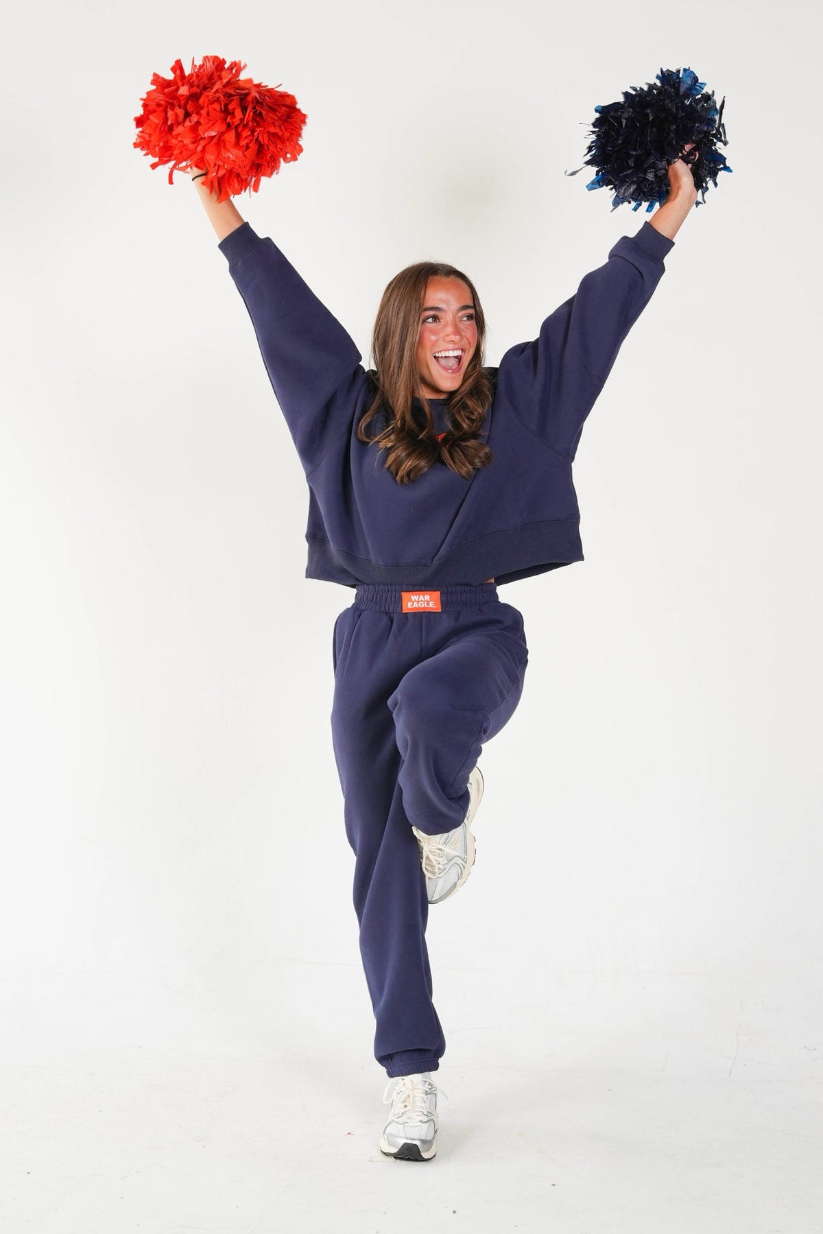 A woman in a navy blue sweatshirt and matching sweatpants jumps while holding red and black pom-poms against a plain white studio background.