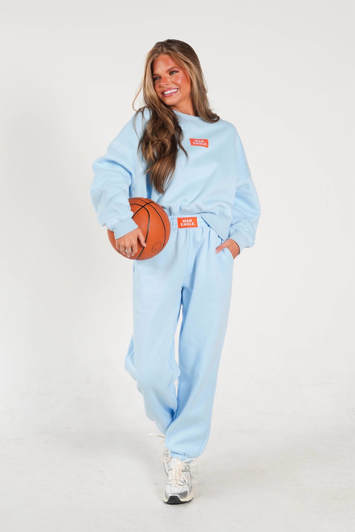 A woman modeling a light blue sweatshirt and matching sweatpants set, holding a basketball. The outfit features a small orange rectangular logo patch on the chest and a matching orange patch at the waist.