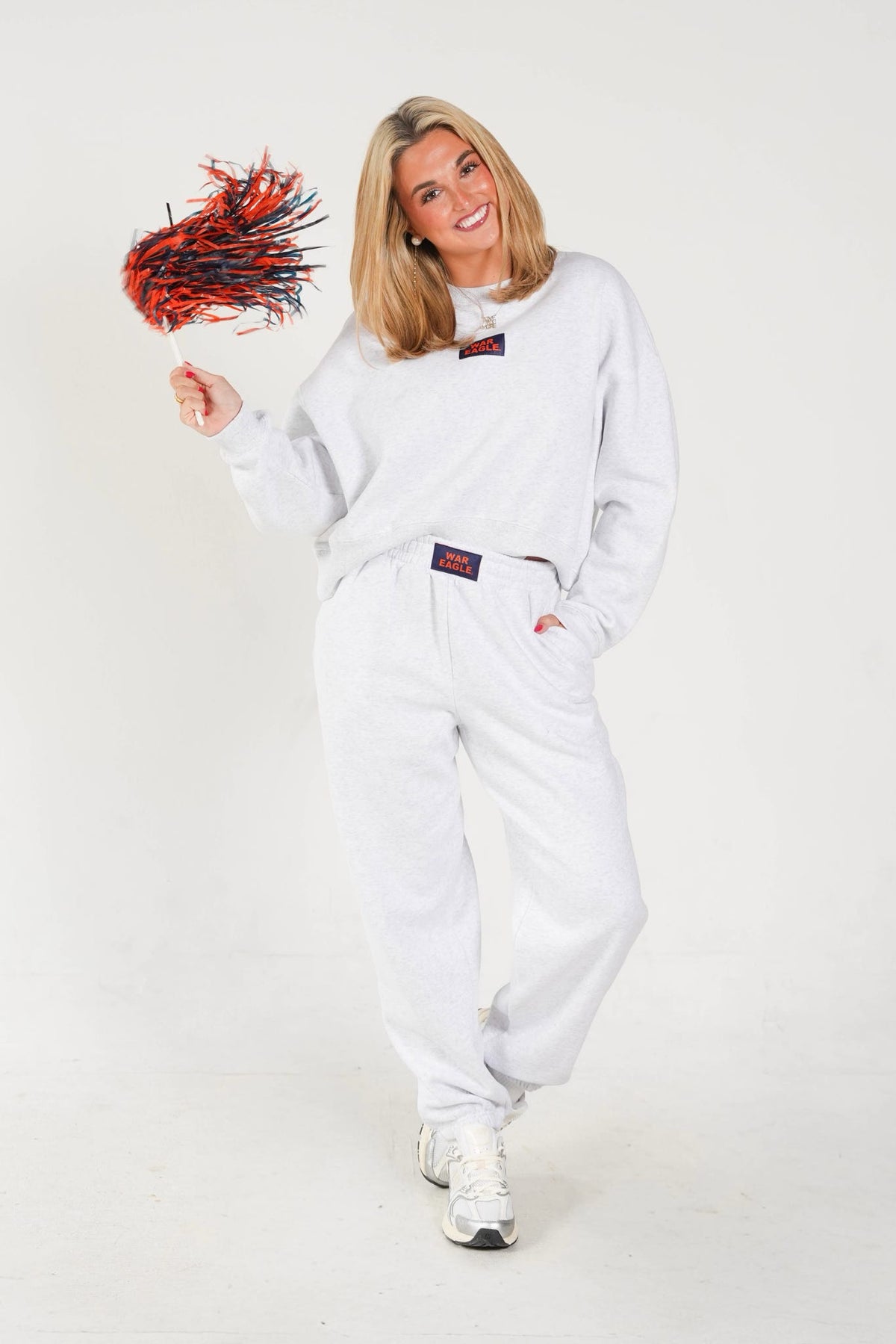 A woman modeling a white sweatshirt and matching sweatpants set, holding a red and black pom-pom against a plain white studio background.