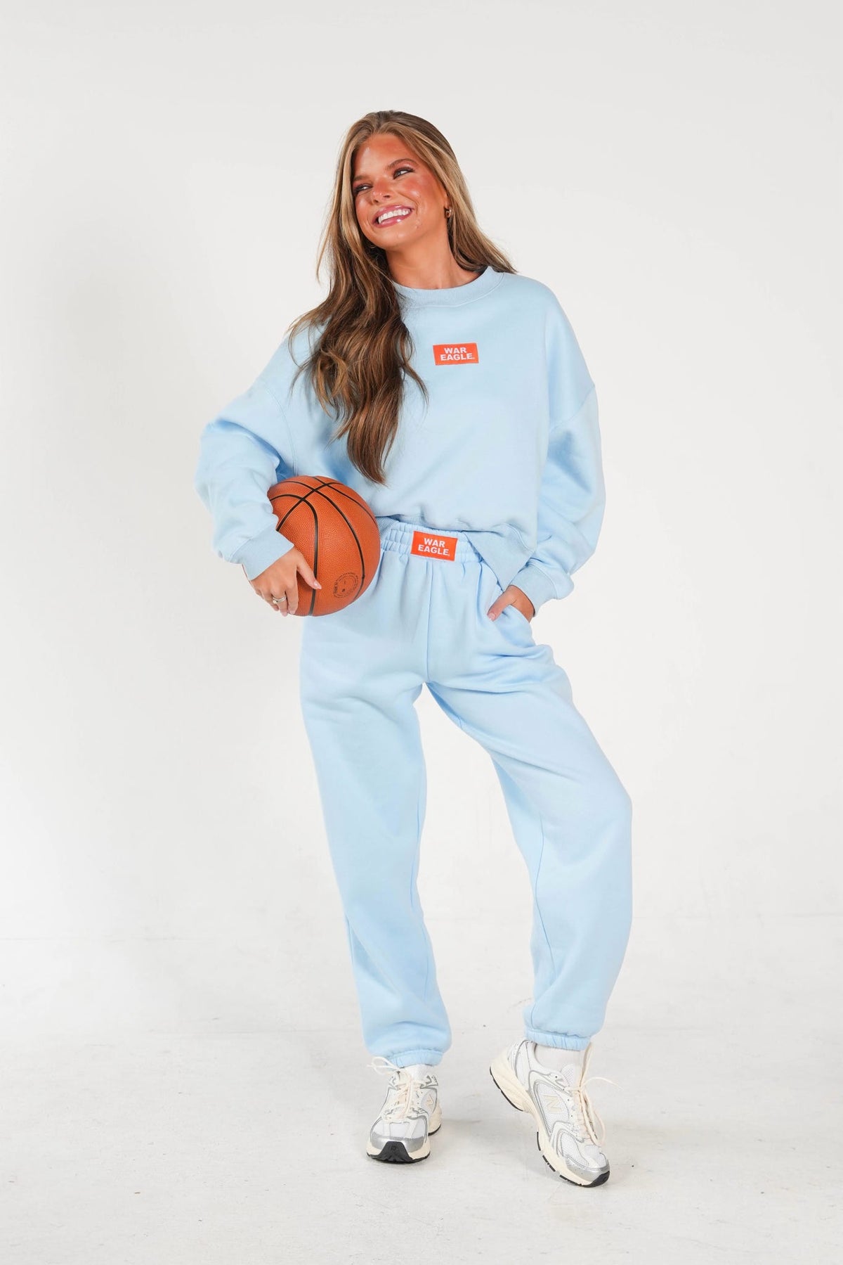 A woman modeling a light blue sweatshirt and sweatpants set with a matching belt and white sneakers, holding a basketball against a plain white studio background.