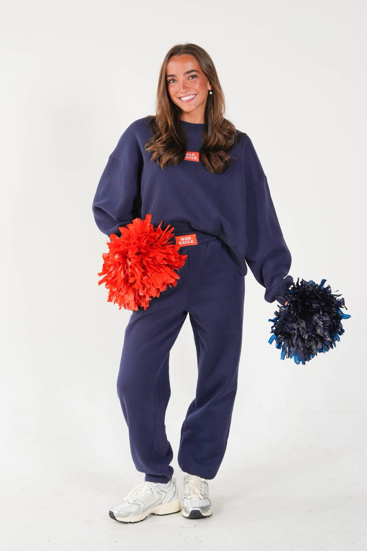 A woman modeling a navy blue sweatshirt and sweatpants set, holding red and navy blue pom-poms. The sweatshirt features a small red rectangular patch on the chest.