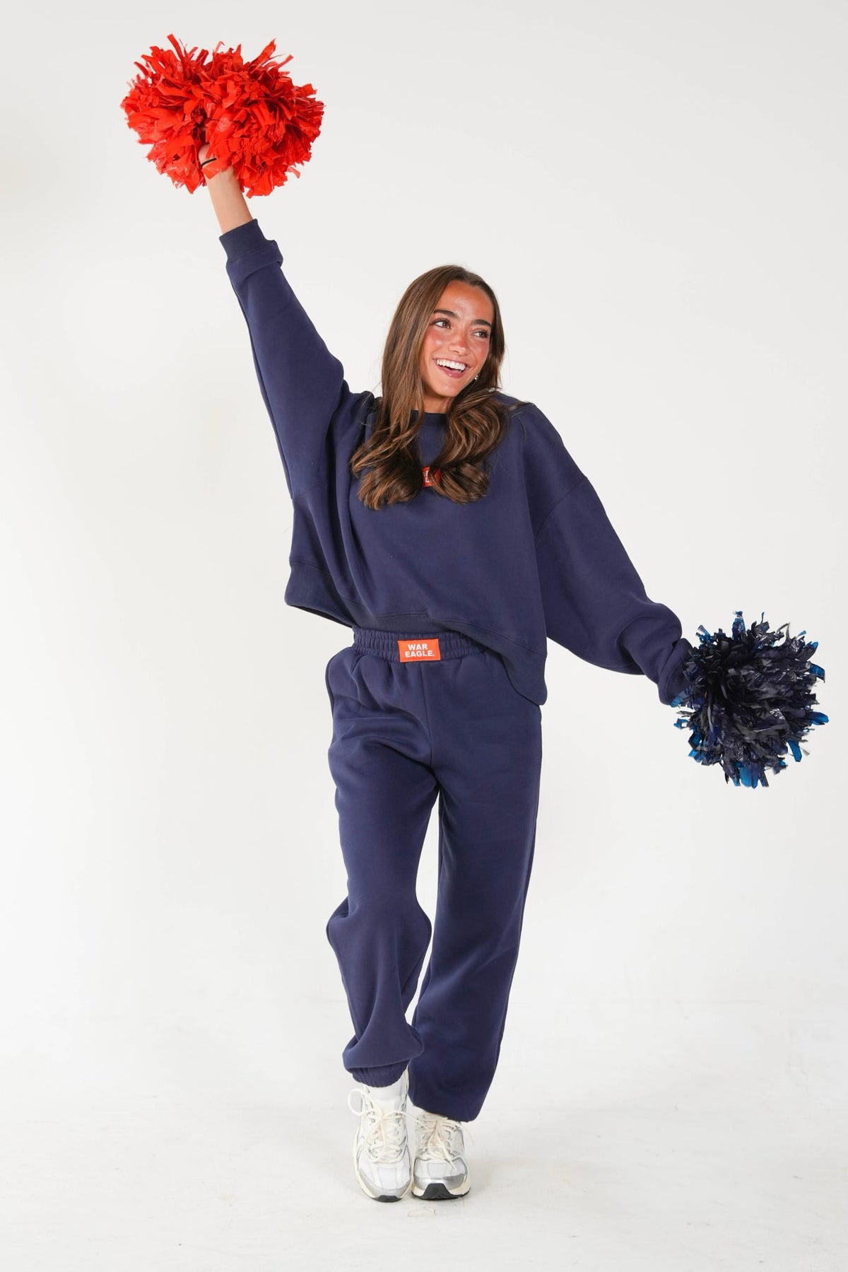 A woman modeling a navy blue sweatshirt and sweatpants set, holding red and blue pom-poms against a plain white studio background.