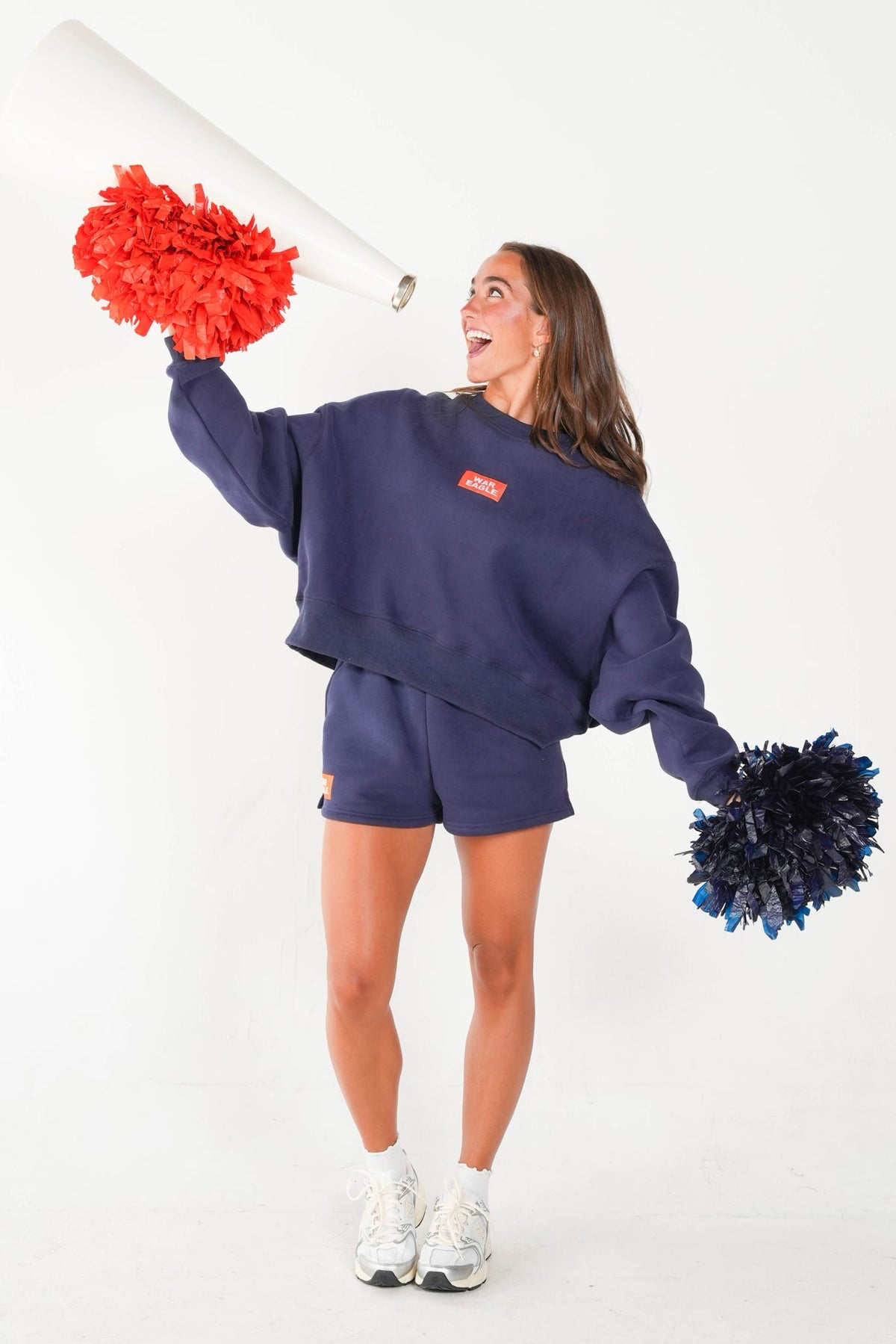 A woman modeling a navy blue cropped sweatshirt and matching shorts, styled like a cheerleader with red and navy pom-poms, a white megaphone, and white sneakers against a plain white studio background.