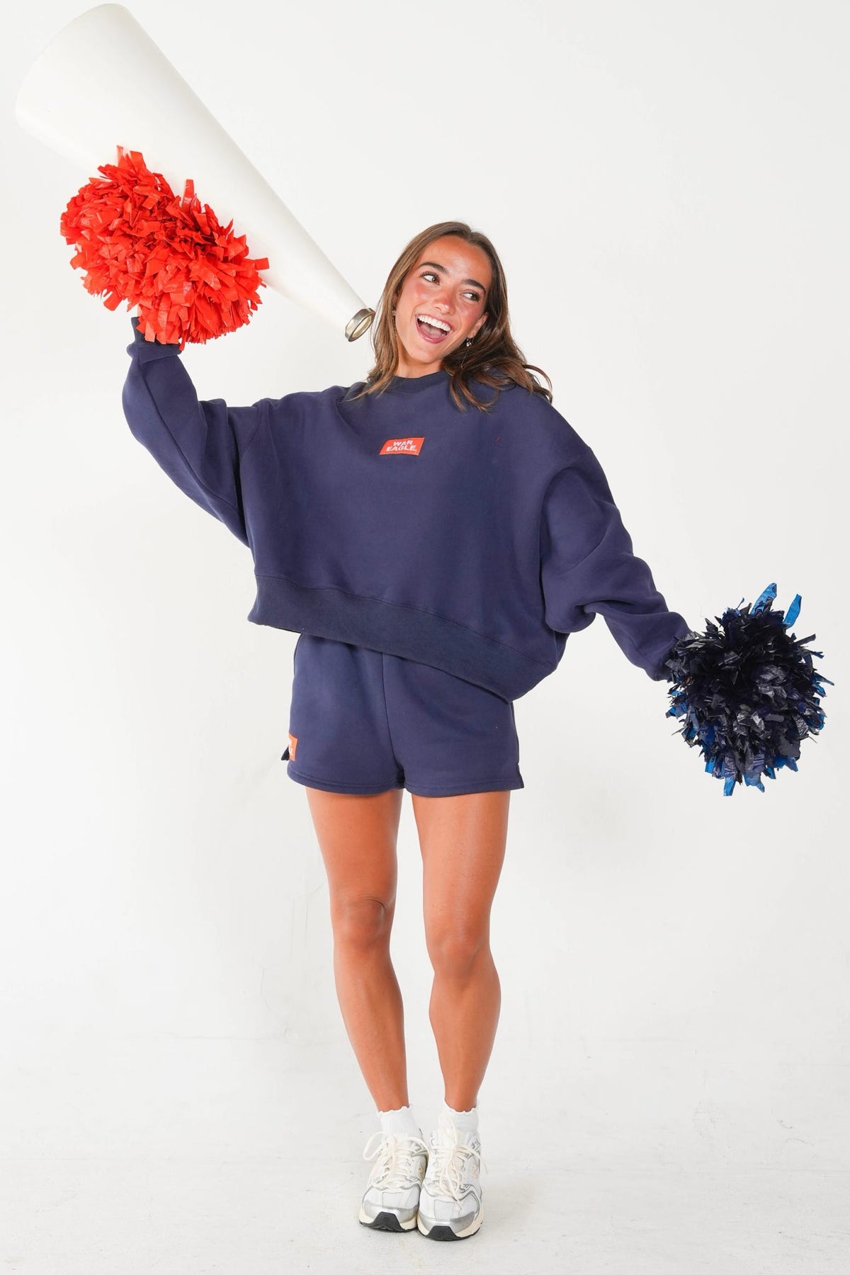 A woman modeling a navy blue sweatshirt and matching shorts set, holding red and blue pom-poms and a white megaphone against a plain white studio background.