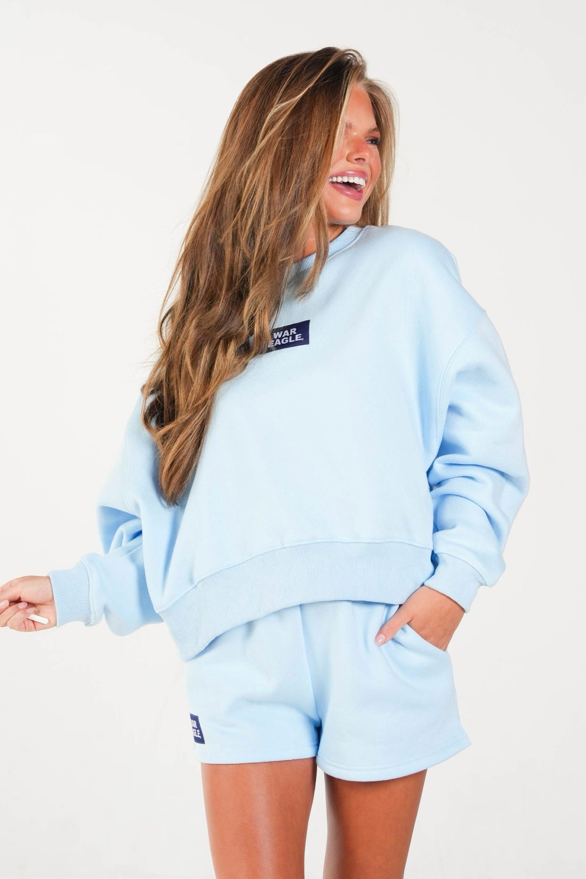 A woman wearing a light blue matching lounge set consisting of a sweatshirt and shorts, with a small dark logo patch on the chest and shorts, photographed against a plain white background.