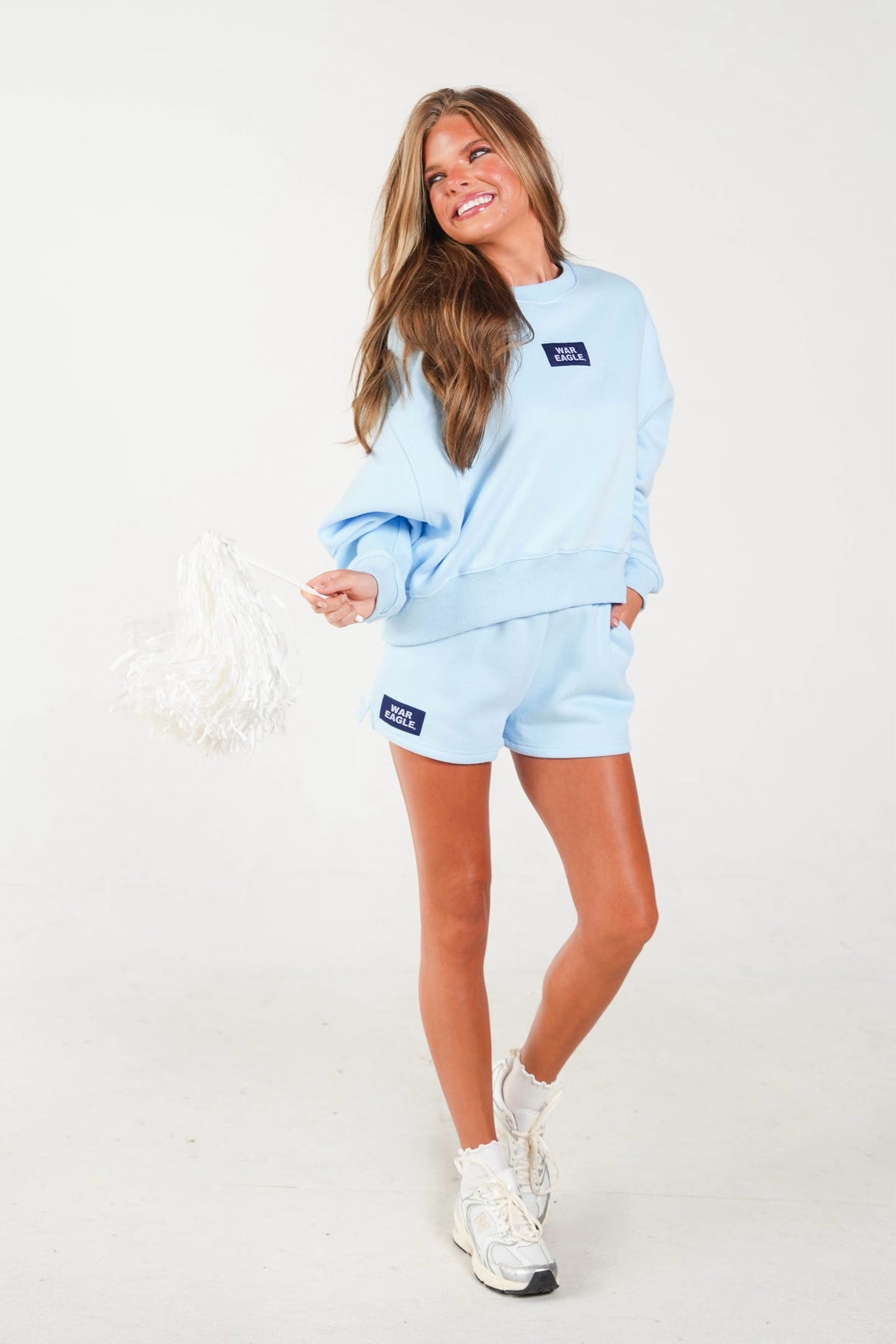 A woman modeling a light blue sweatshirt and matching shorts set, featuring a small dark blue rectangular logo patch on the chest and left leg. She is holding a white pom-pom and wearing white sneakers against a plain white studio background.