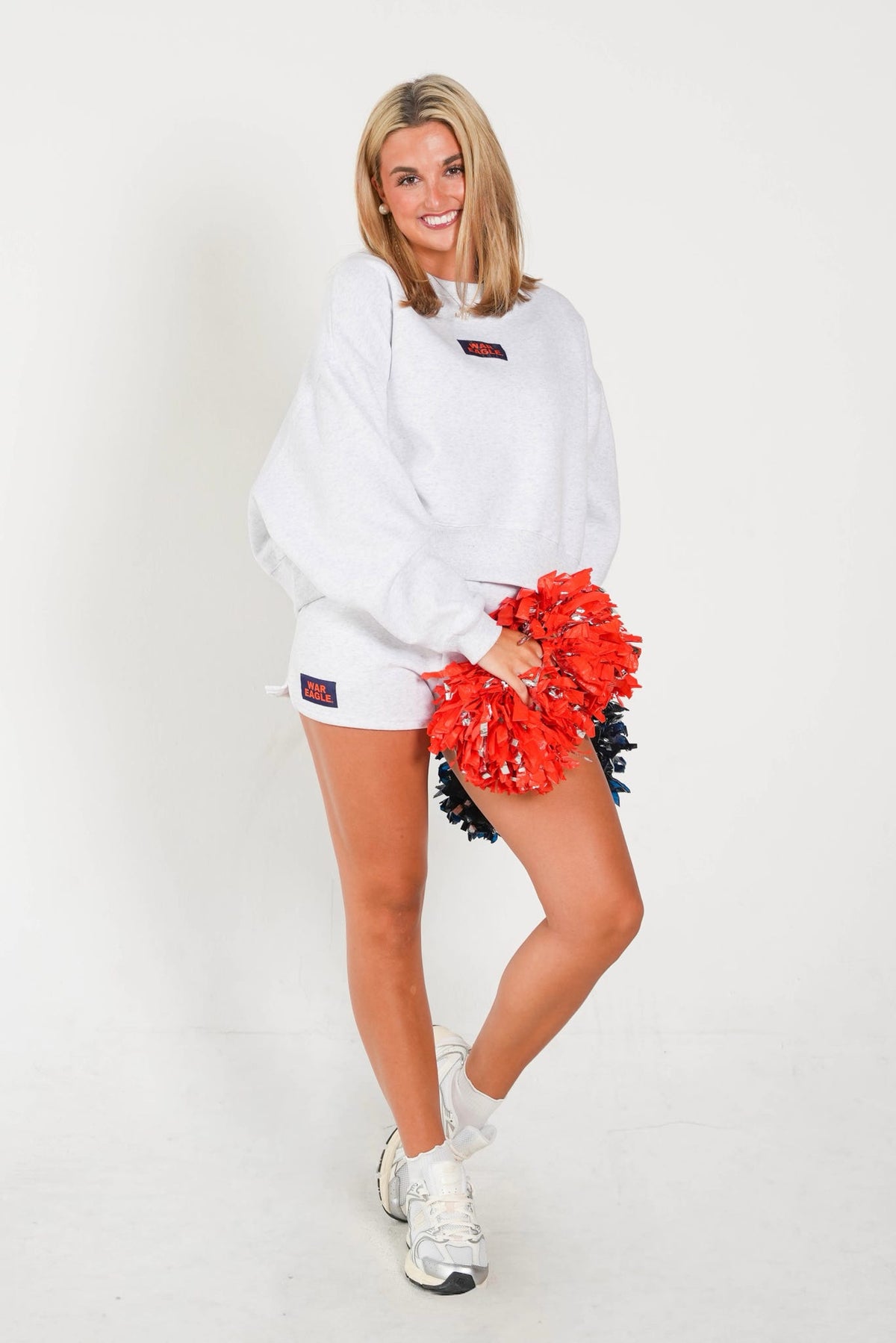 A woman modeling a white oversized sweatshirt and matching white shorts, holding a red and black pom-pom. She is standing against a plain white studio background.