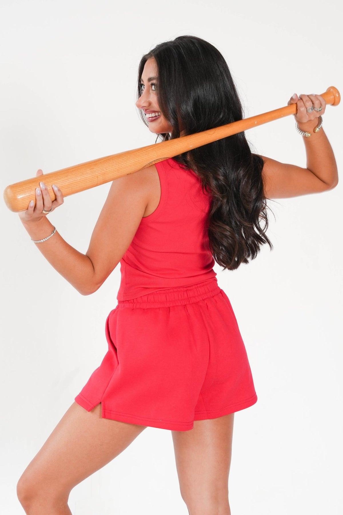 A woman with long dark hair wearing a bright red sleeveless top and matching red shorts, holding a wooden baseball bat over her shoulder against a plain white background.