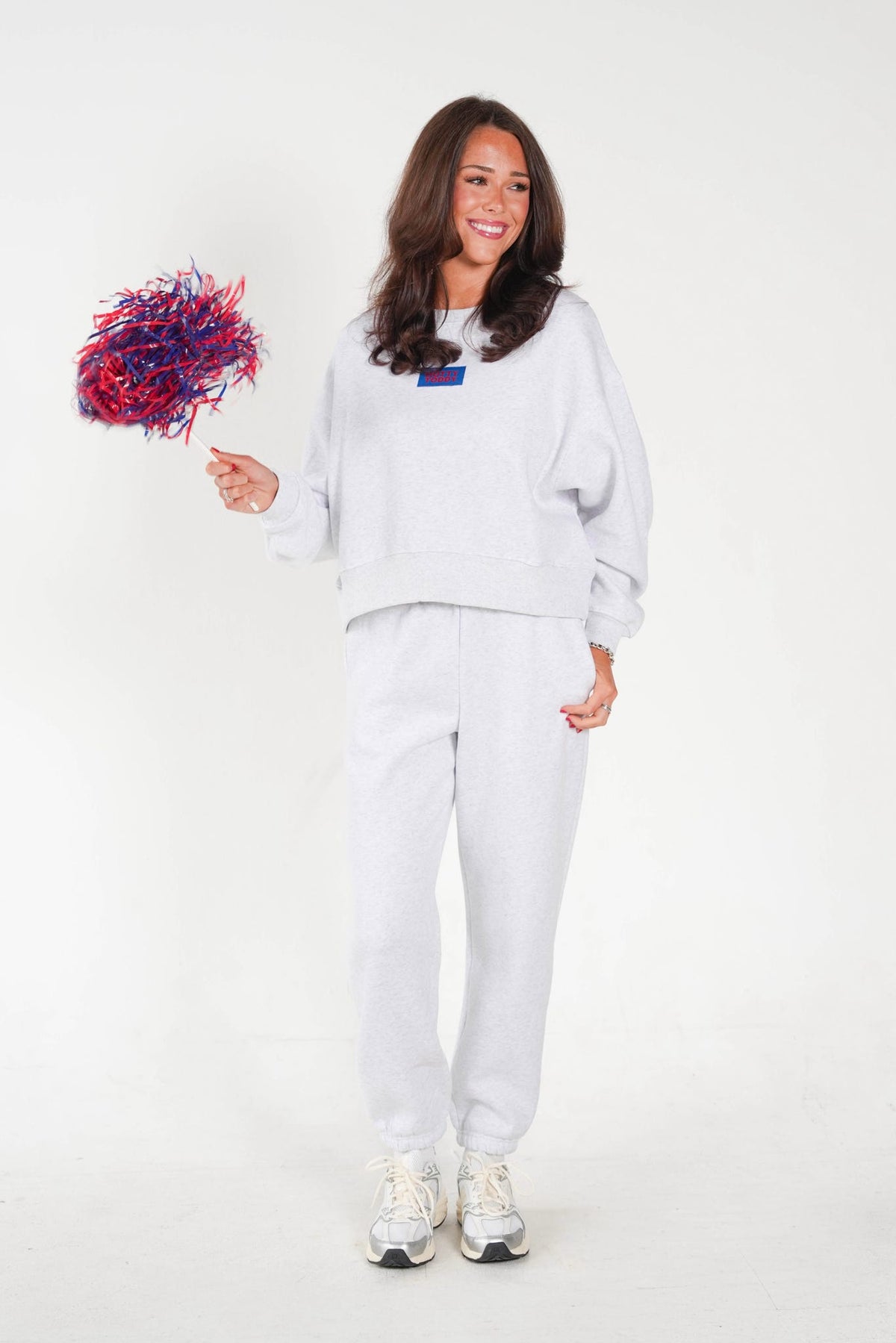 A woman modeling a light gray sweatshirt and sweatpants set, holding a red and blue pom-pom against a plain white studio background.