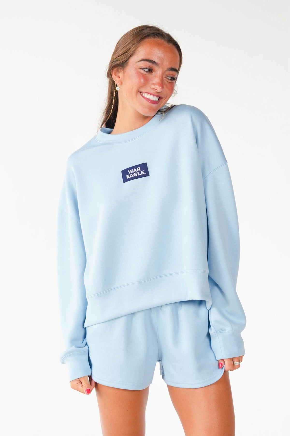 A woman modeling a light blue sweatshirt and matching shorts set. The sweatshirt features a small dark blue rectangular chest patch with the text 'MAN BACK' in white.