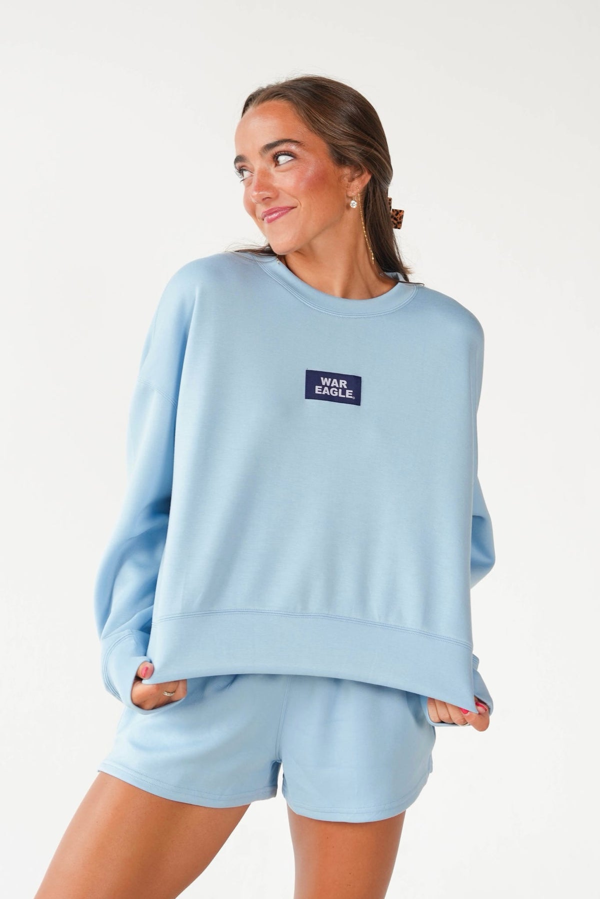 A woman wearing a light blue sweatshirt and matching shorts set, featuring a small dark navy rectangular chest patch with the text 'WAN EXCILE.' in white, photographed against a clean white studio background.