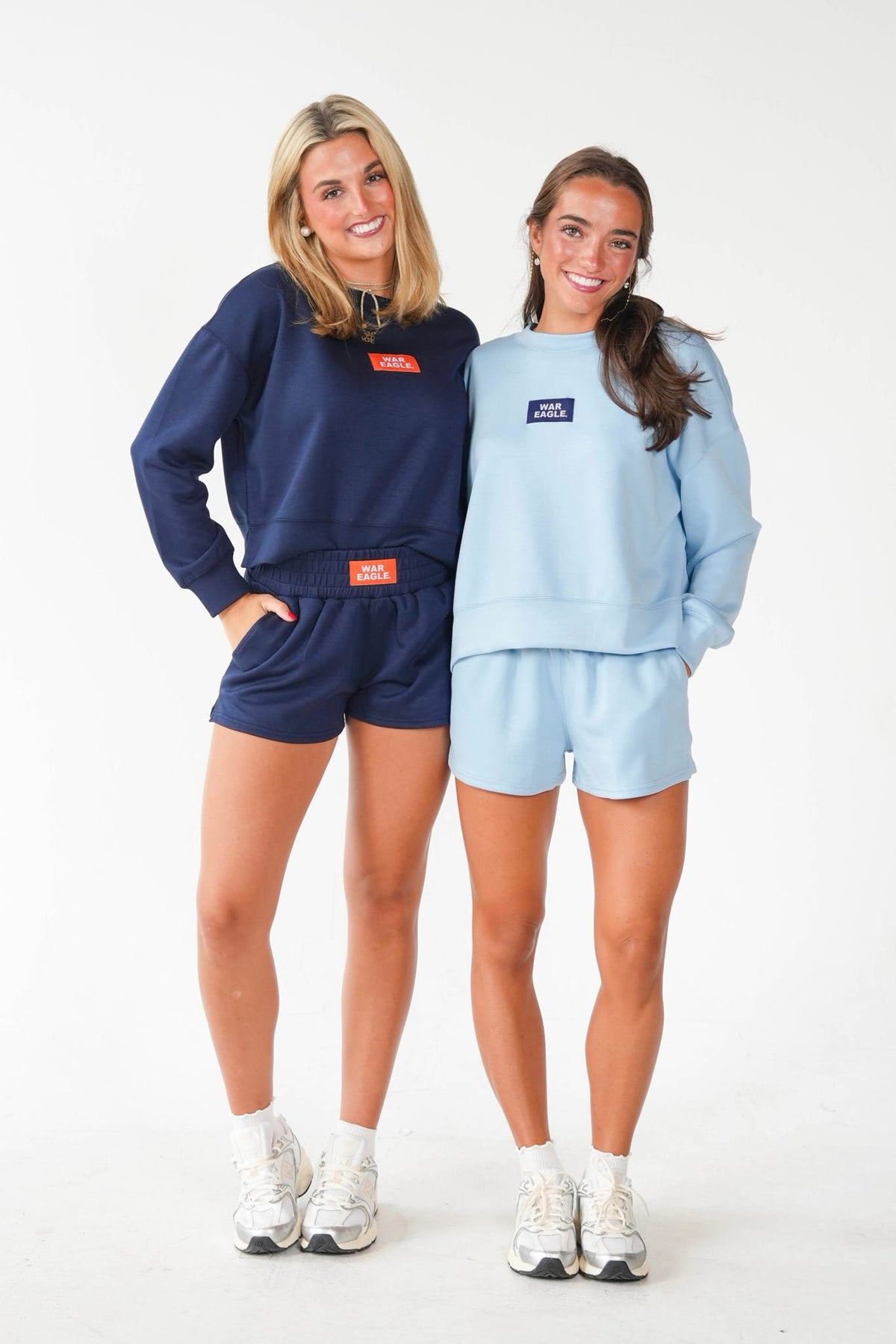 Two women modeling matching athletic sweatshirt and shorts sets in a studio setting. The woman on the left wears a dark navy blue set with a small red rectangular logo patch on the chest and waist.