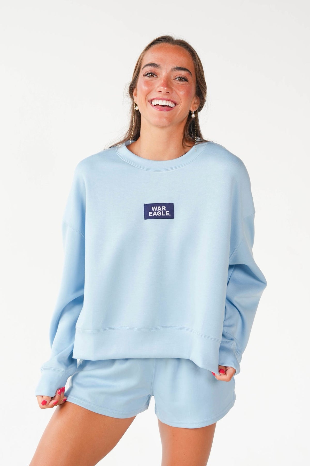 A woman modeling a light blue sweatshirt and matching shorts set. The sweatshirt features a small dark blue rectangular chest patch with the text 'WAR EAGLE' in white.