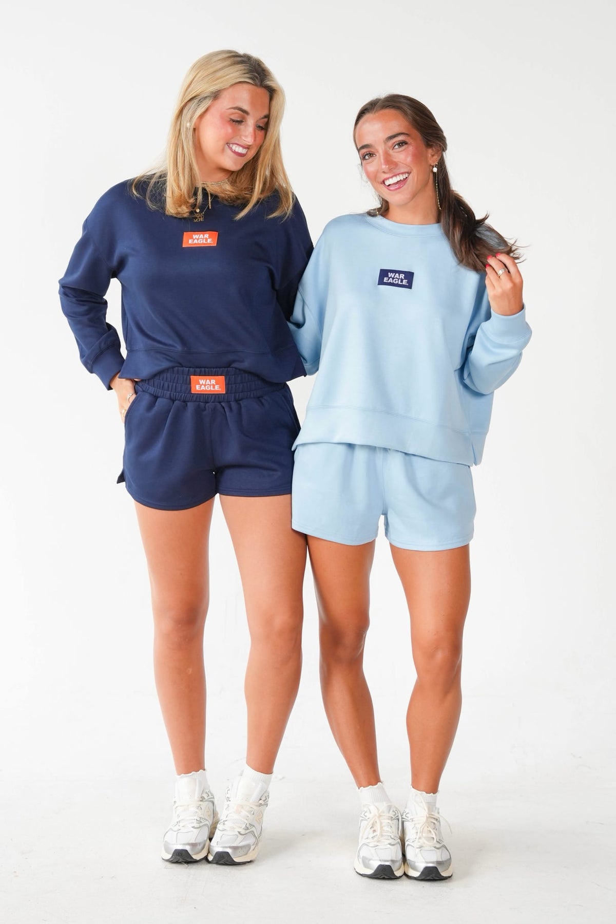 Two women modeling matching lounge sets in a studio setting. The woman on the left wears a navy blue sweatshirt and shorts set with an orange logo patch, and the woman on the right wears a light blue sweatshirt and shorts set with a navy logo patch.