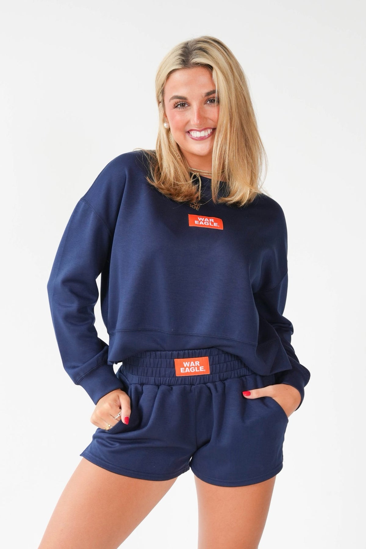A woman modeling a navy blue sweatshirt and matching shorts set. The set features a small orange rectangular patch on the chest with 'WAR EAGLE' printed in white, and a larger matching patch on the elastic waistband.