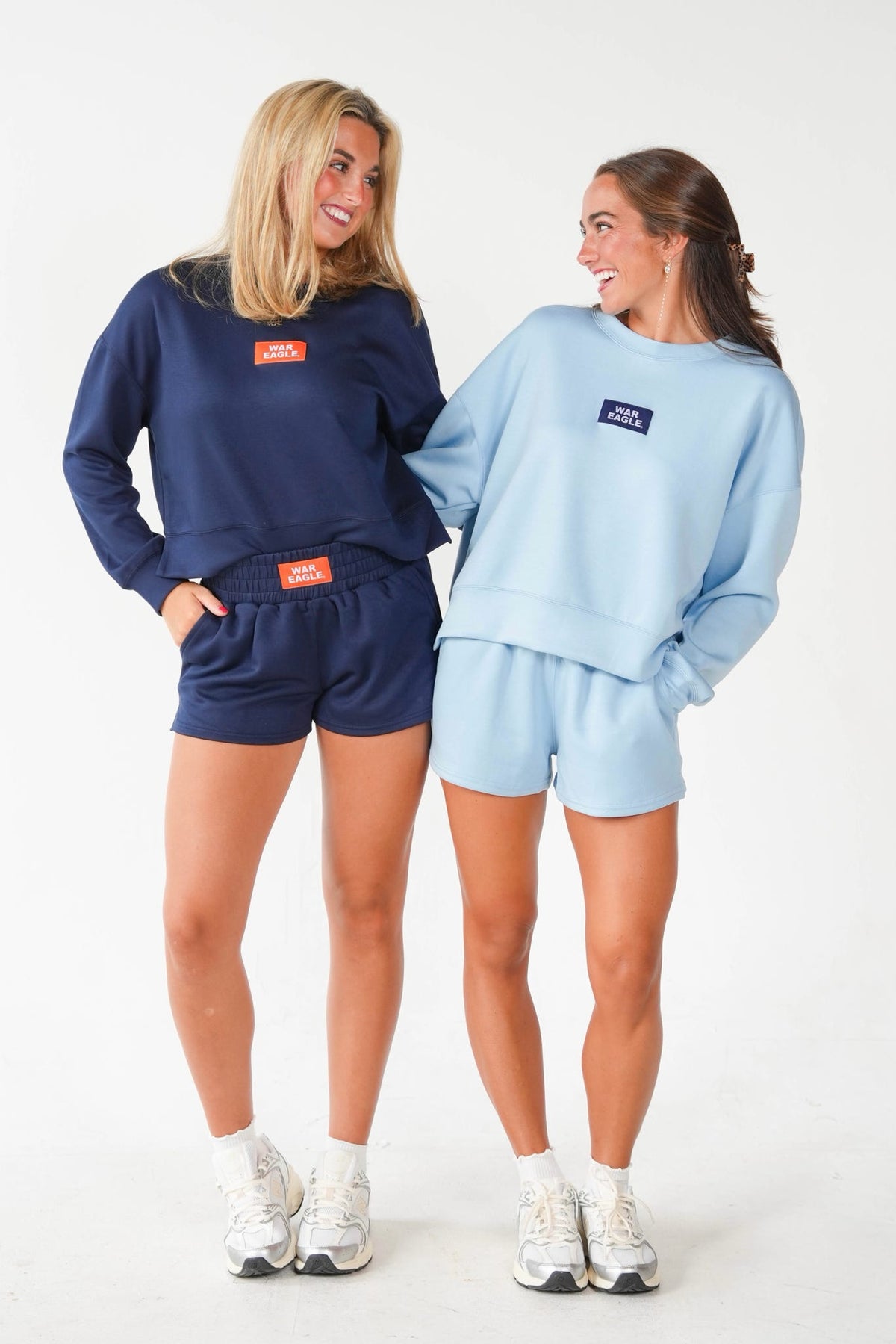 Two women modeling matching two-piece lounge sets in different colors: one in navy blue and one in light blue. Each set includes a long-sleeve sweatshirt and shorts with a small rectangular chest logo patch.