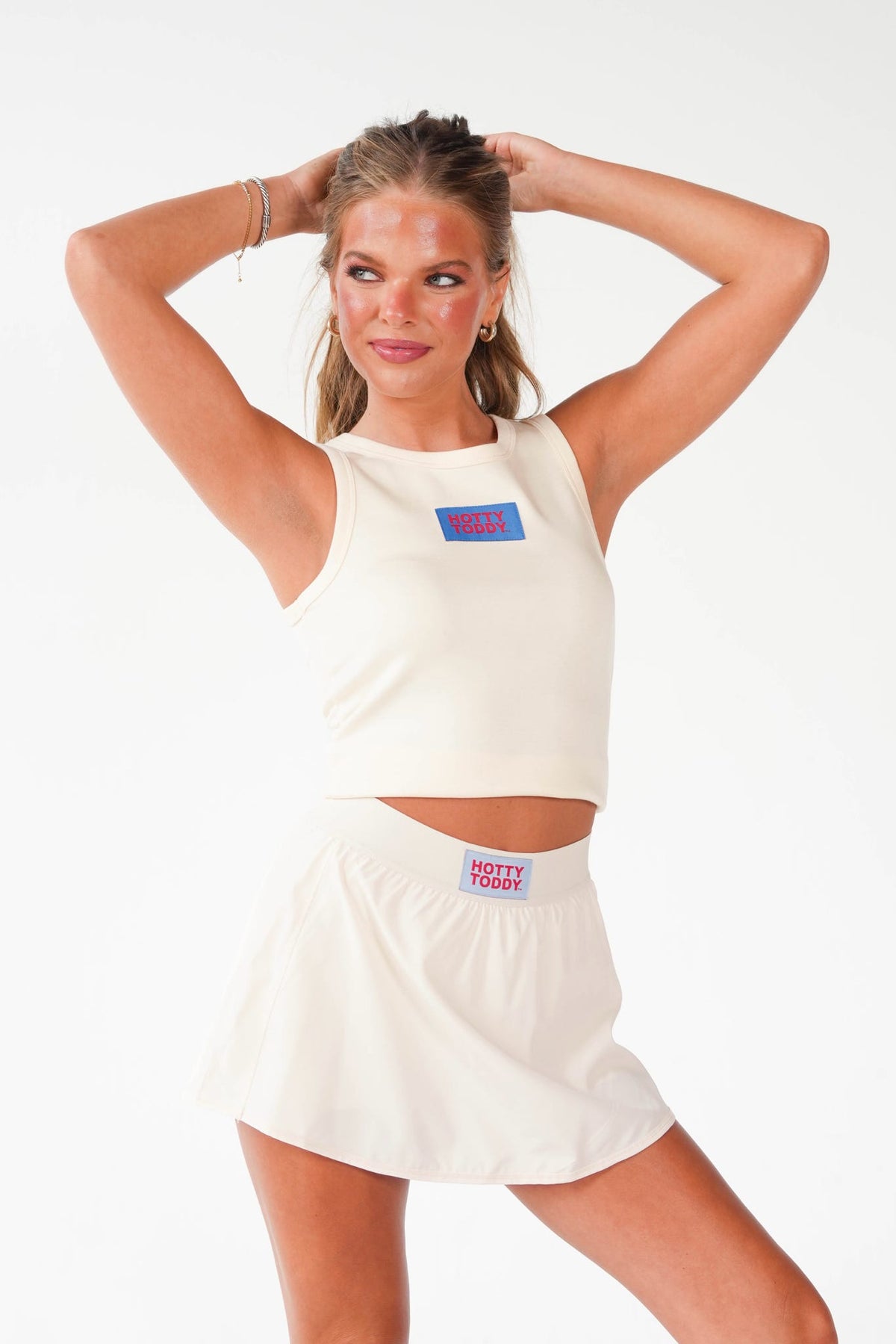 A woman modeling a matching off-white athletic set consisting of a cropped tank top and a high-waisted mini skirt, both featuring a small colorful 'HOSTELITY' brand patch on the chest and waistband.
