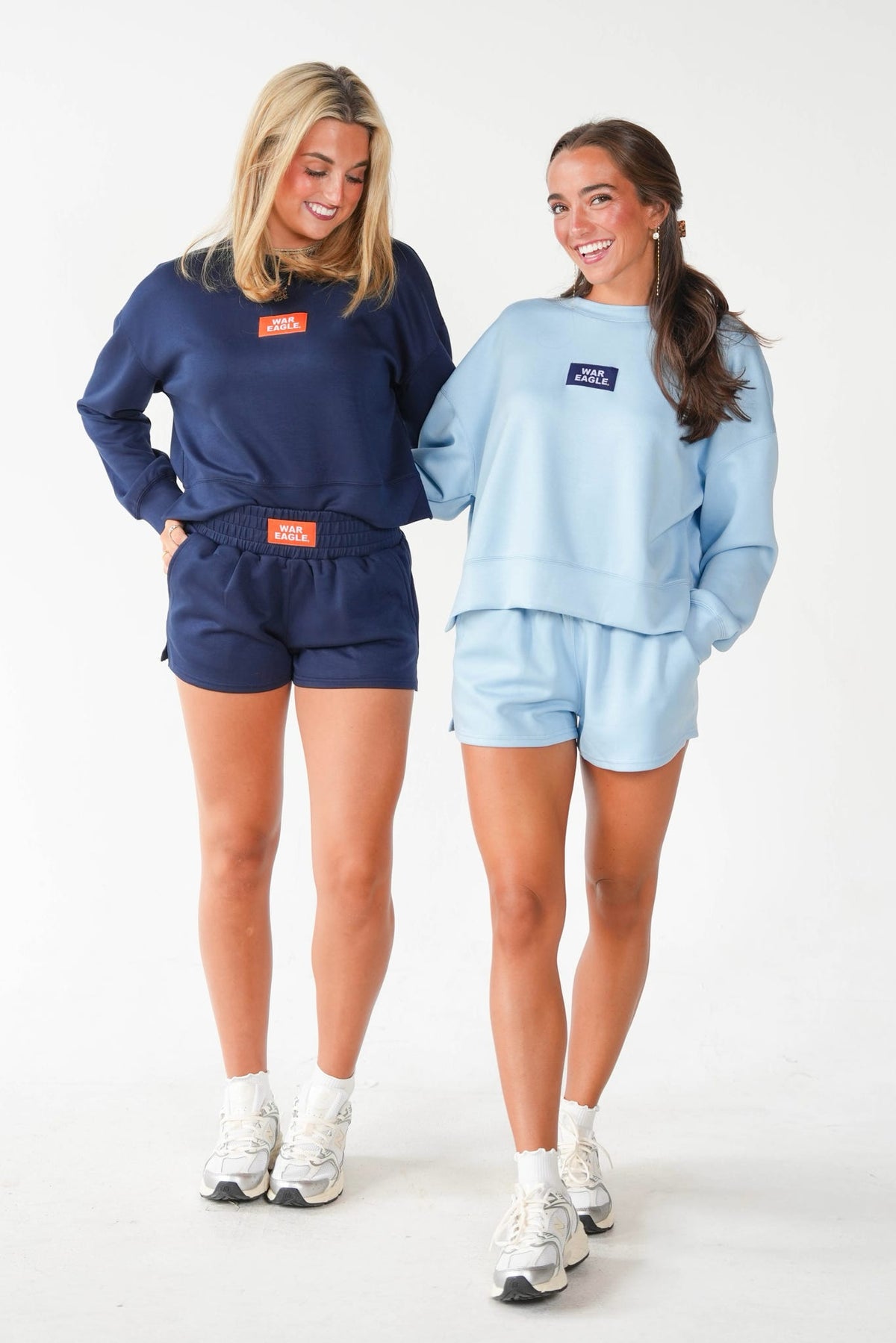 Two women modeling matching sweatshirt and shorts sets in a studio setting. The woman on the left wears a dark navy blue set with an orange rectangular chest patch, and the woman on the right wears a light blue set with a dark blue rectangular chest patch.